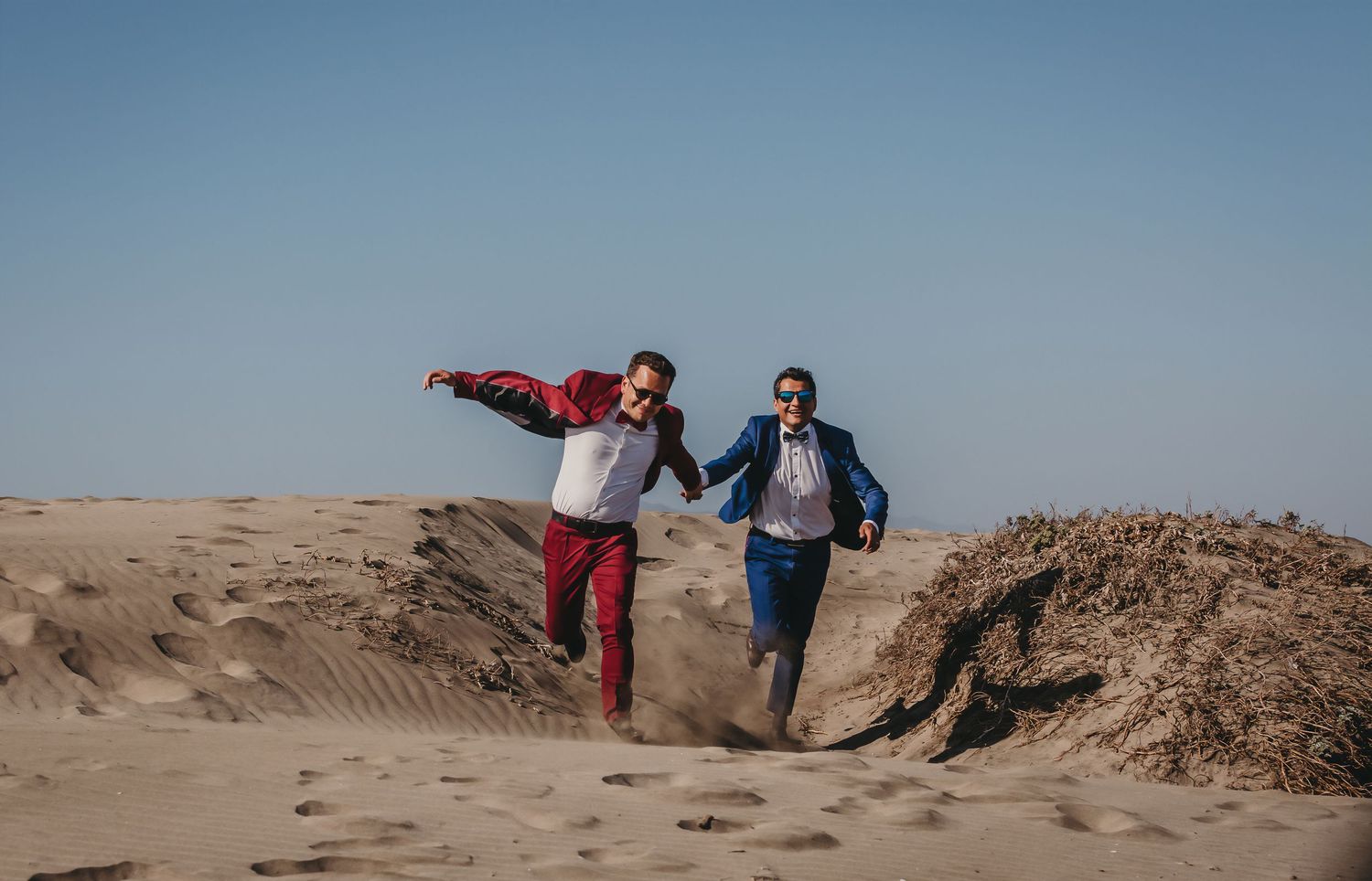 Two grooms in red and blue suits running joyfully across sandy desert dunes on sunny day.