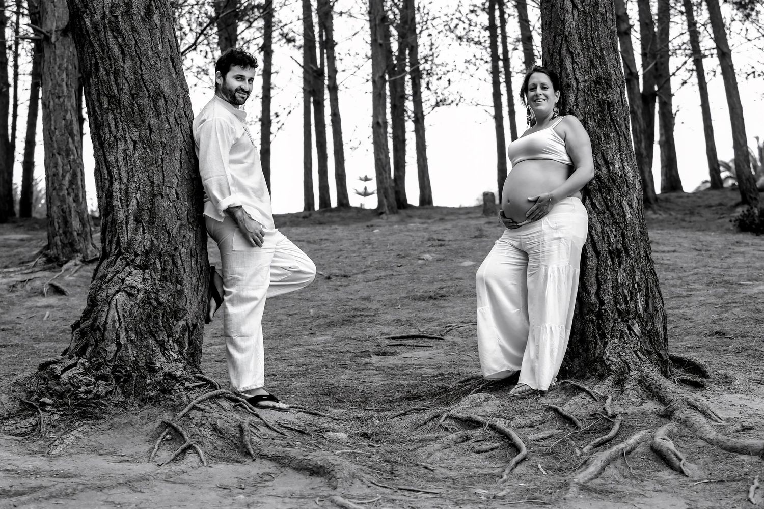 Black and white maternity photo of pregnant couple standing by tree roots in a forest, wearing white outfits.