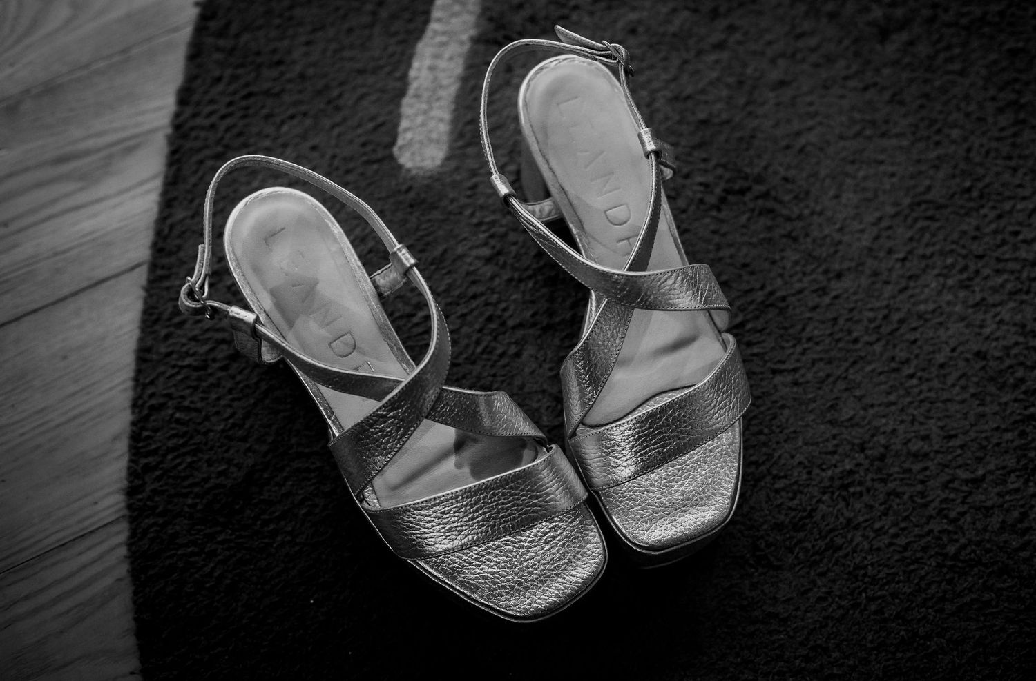 Black and white photo of strappy heeled sandals with ankle buckles resting on a textured carpet floor.