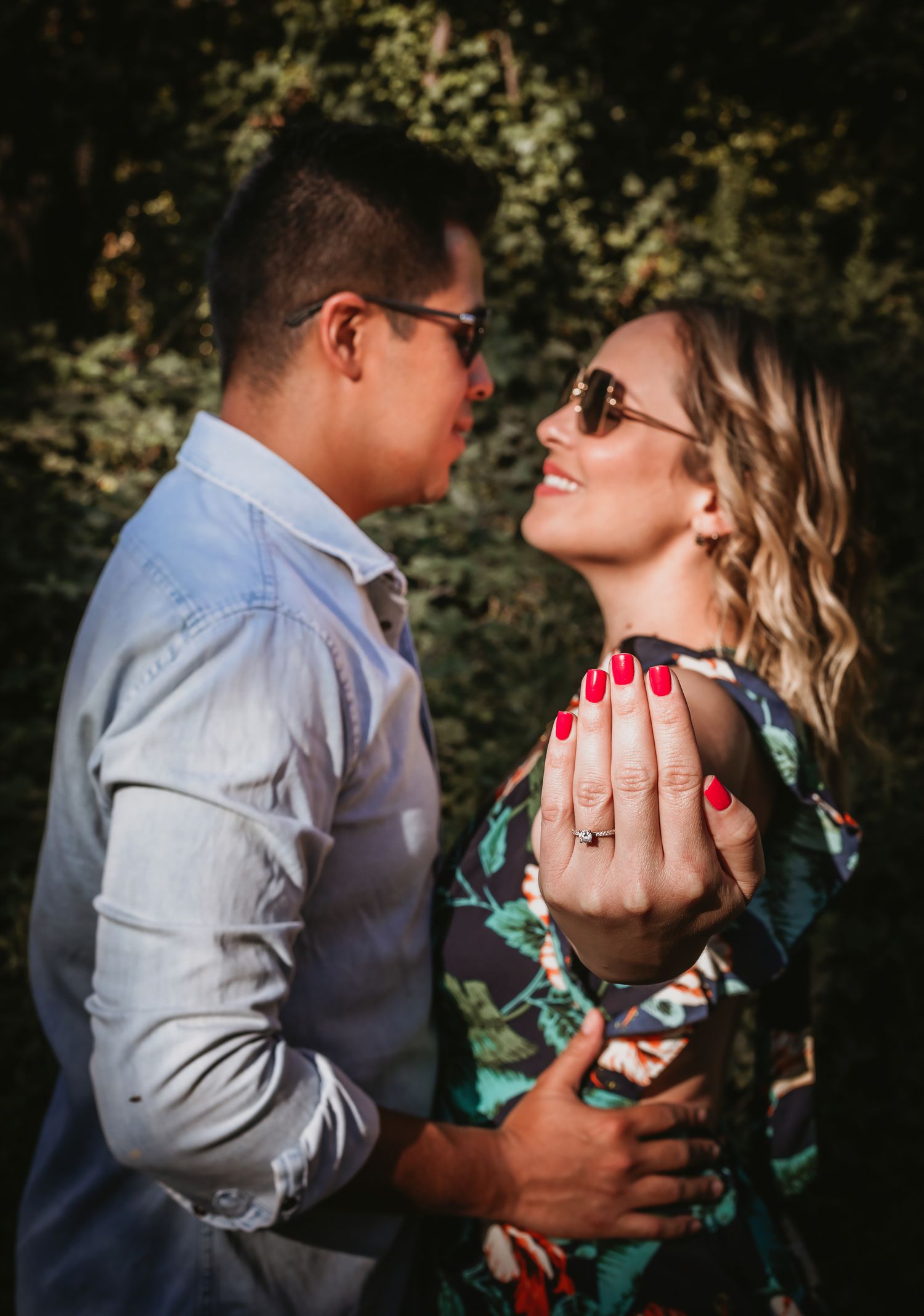 Couple gazing at each other outdoors, woman showing engagement ring with red nails, both wearing sunglasses.