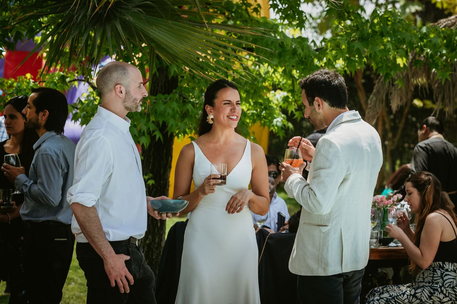 Bride in white dress laughing with guests at outdoor wedding reception surrounded by lush greenery.