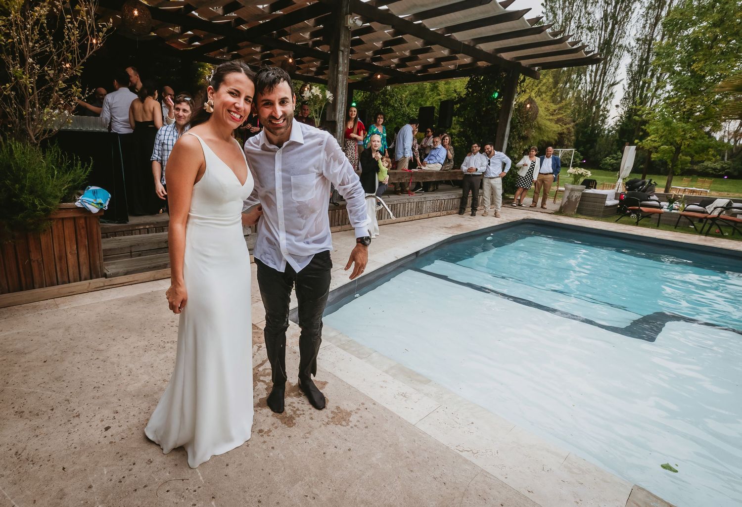Bride in white gown and groom in casual attire pose smiling by outdoor pool at wedding reception.