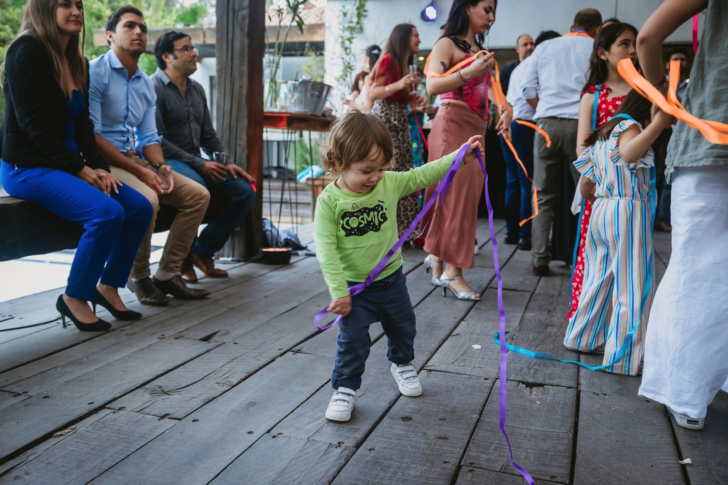 Toddler playing with colorful ribbons on wooden floor at a festive indoor party with guests seated nearby.