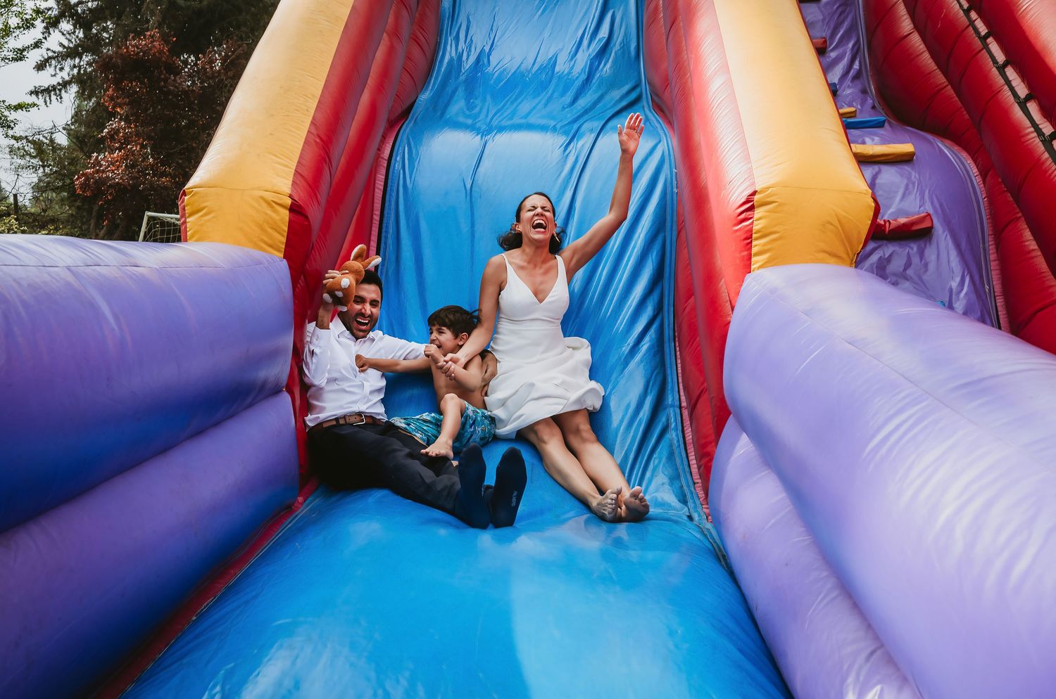 Bride and groom laughing on colorful inflatable bounce house slide at outdoor wedding reception.