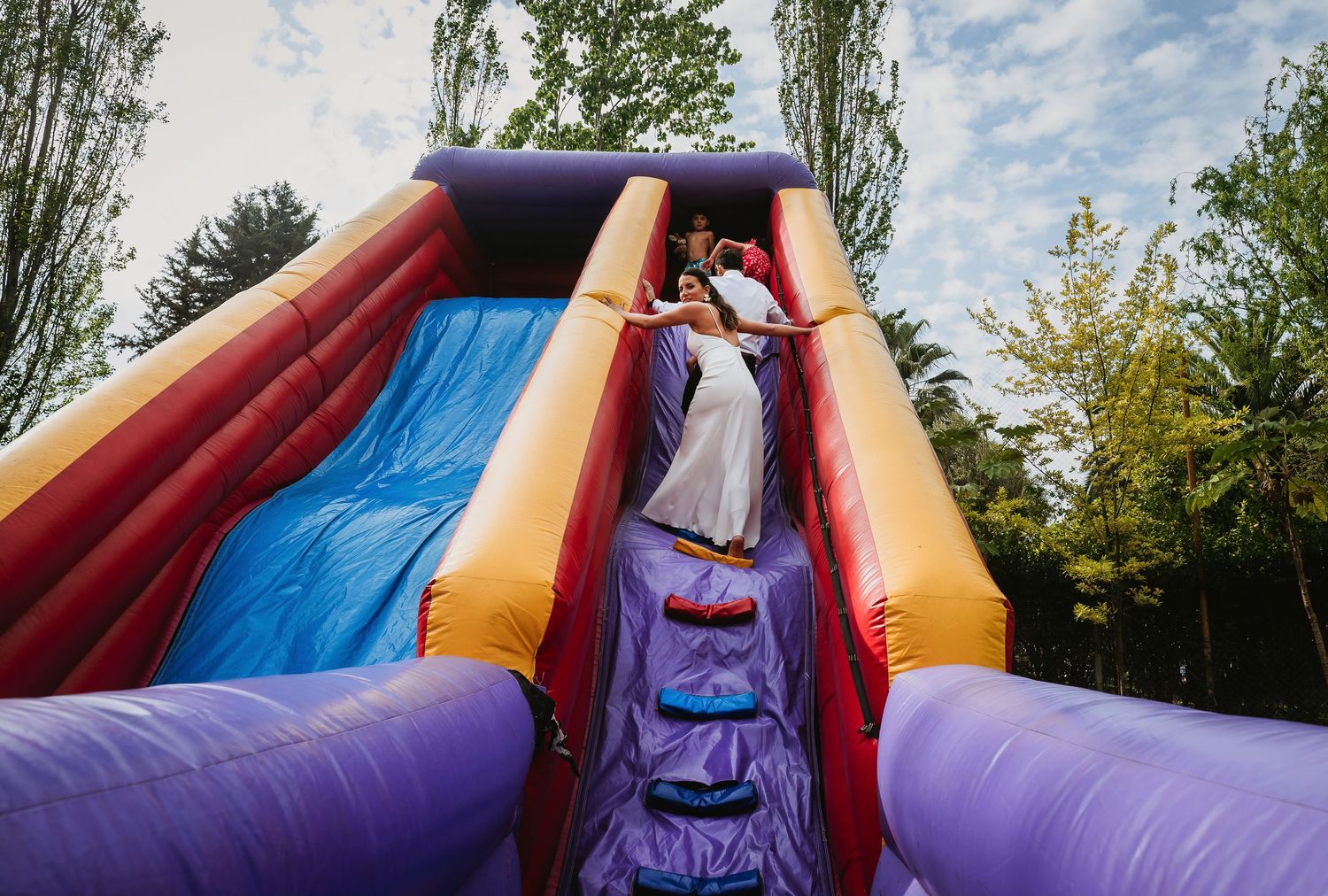 Bride and groom sliding down colorful inflatable bounce house slide at outdoor wedding celebration.