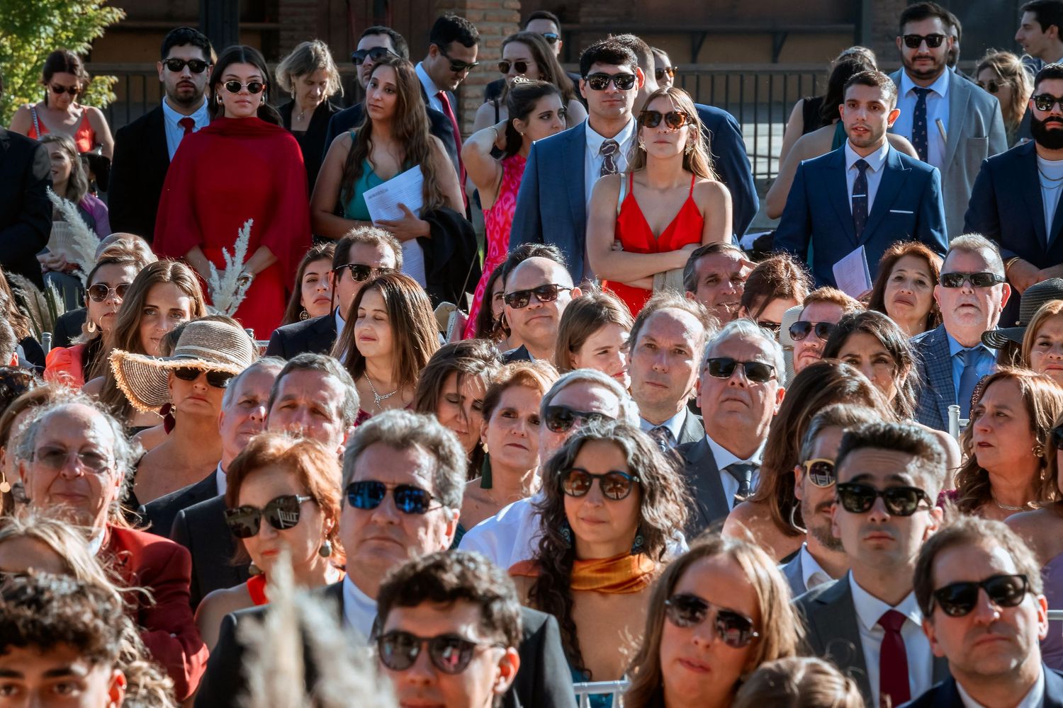 Large crowd of elegantly dressed wedding guests seated at outdoor ceremony.