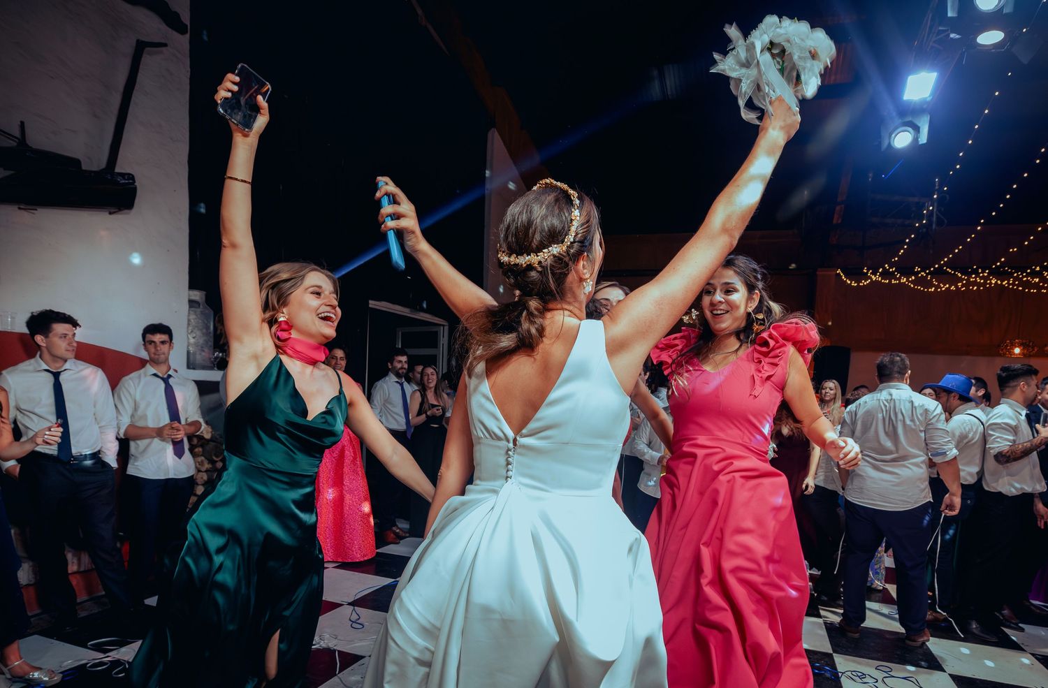 Bride raises bouquet on dance floor flanked by colorful bridesmaids at wedding.