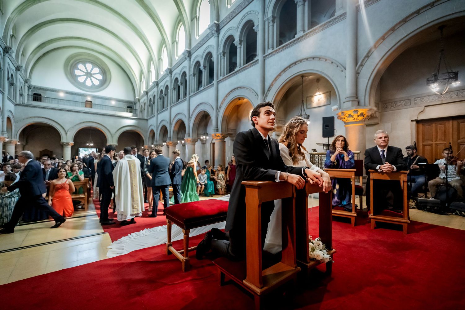 Bride and groom kneeling at red-carpeted altar in ornate church with wedding guests seated in background.