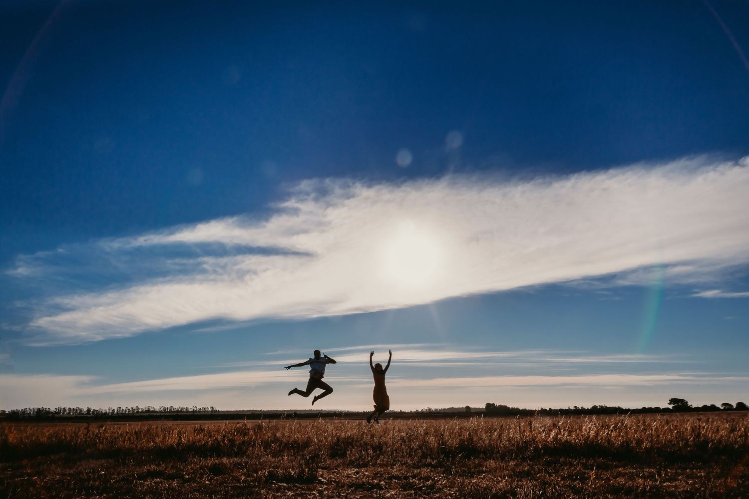Two people jumping joyfully in an open field against a vivid blue sky with scattered clouds.
