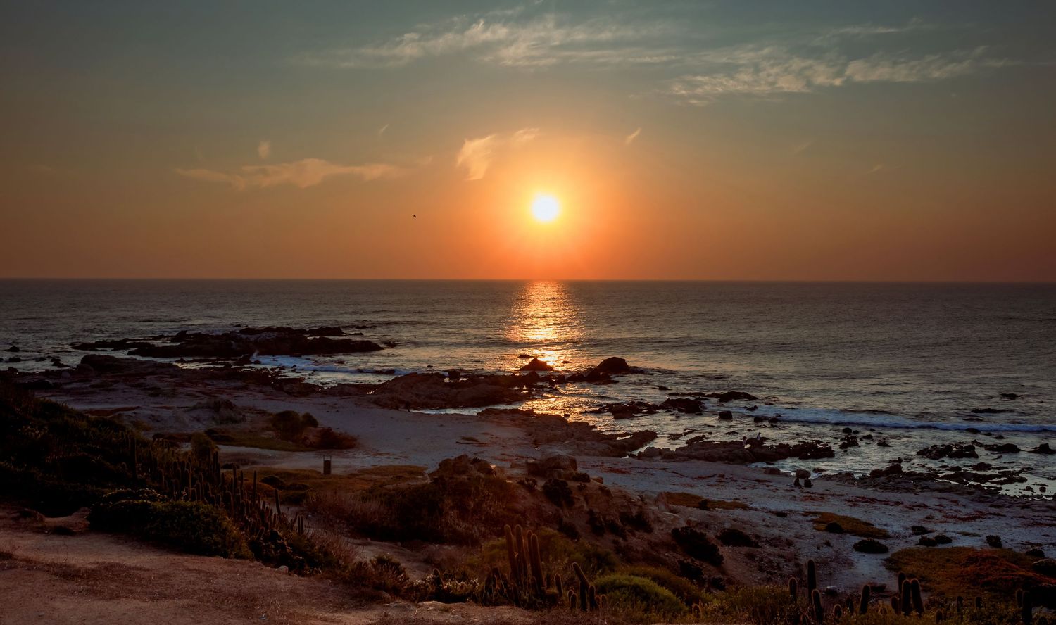 Stunning coastal sunset with golden orange sky reflecting on calm ocean waters over rocky shoreline.