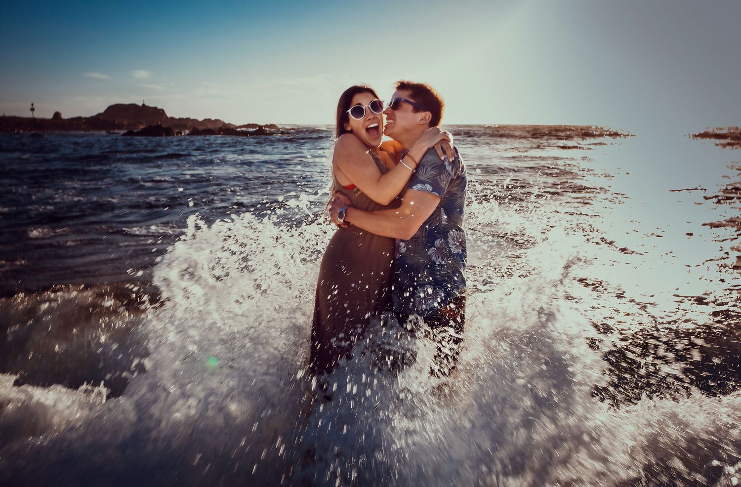 Couple hugging joyfully as ocean waves splash around them on a sunny beach.