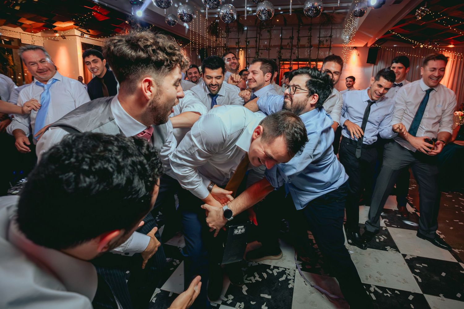 Groomsmen huddled together in energetic group moment on wedding reception dance floor.