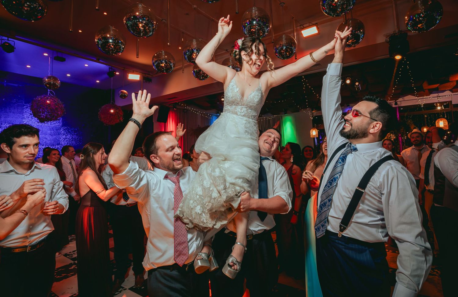 Bride lifted by guests on lively wedding dance floor with disco balls and colorful lights.