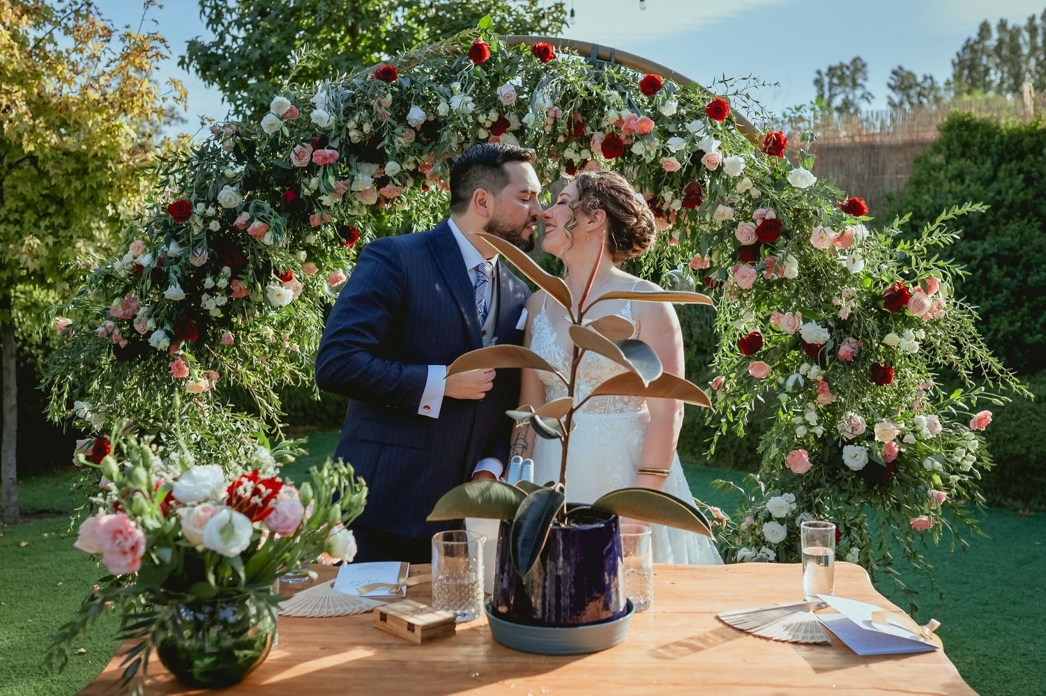 Bride and groom kiss romantically beneath floral arch at outdoor garden wedding ceremony table.