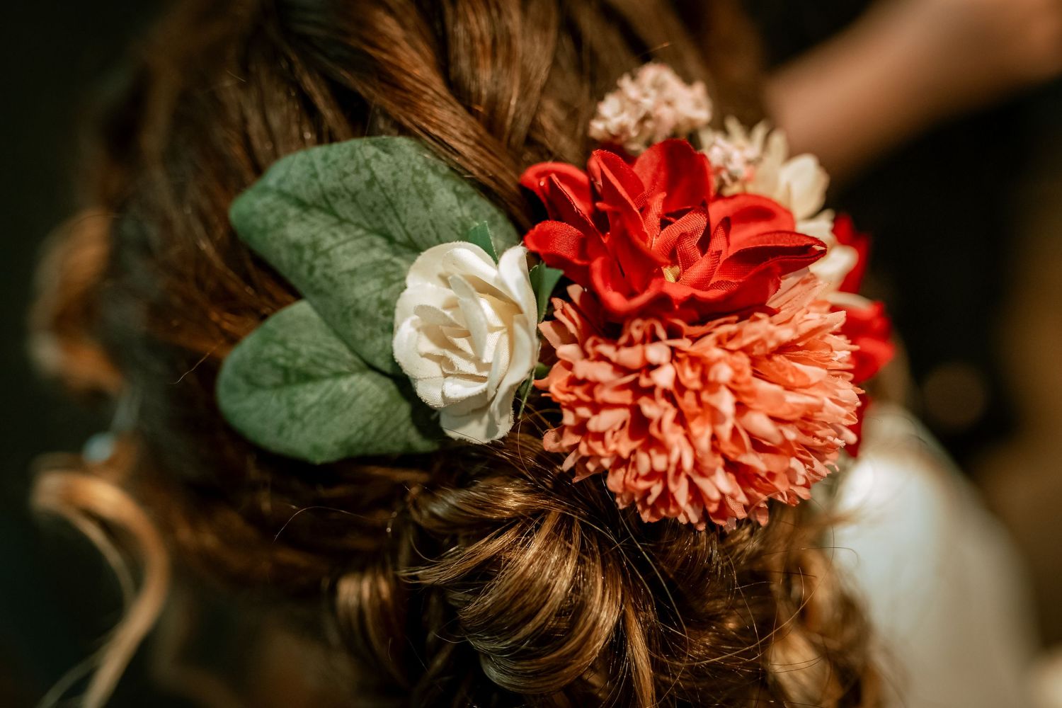 Close-up of bridal updo hairstyle adorned with red, coral, and white flowers with green leaves.