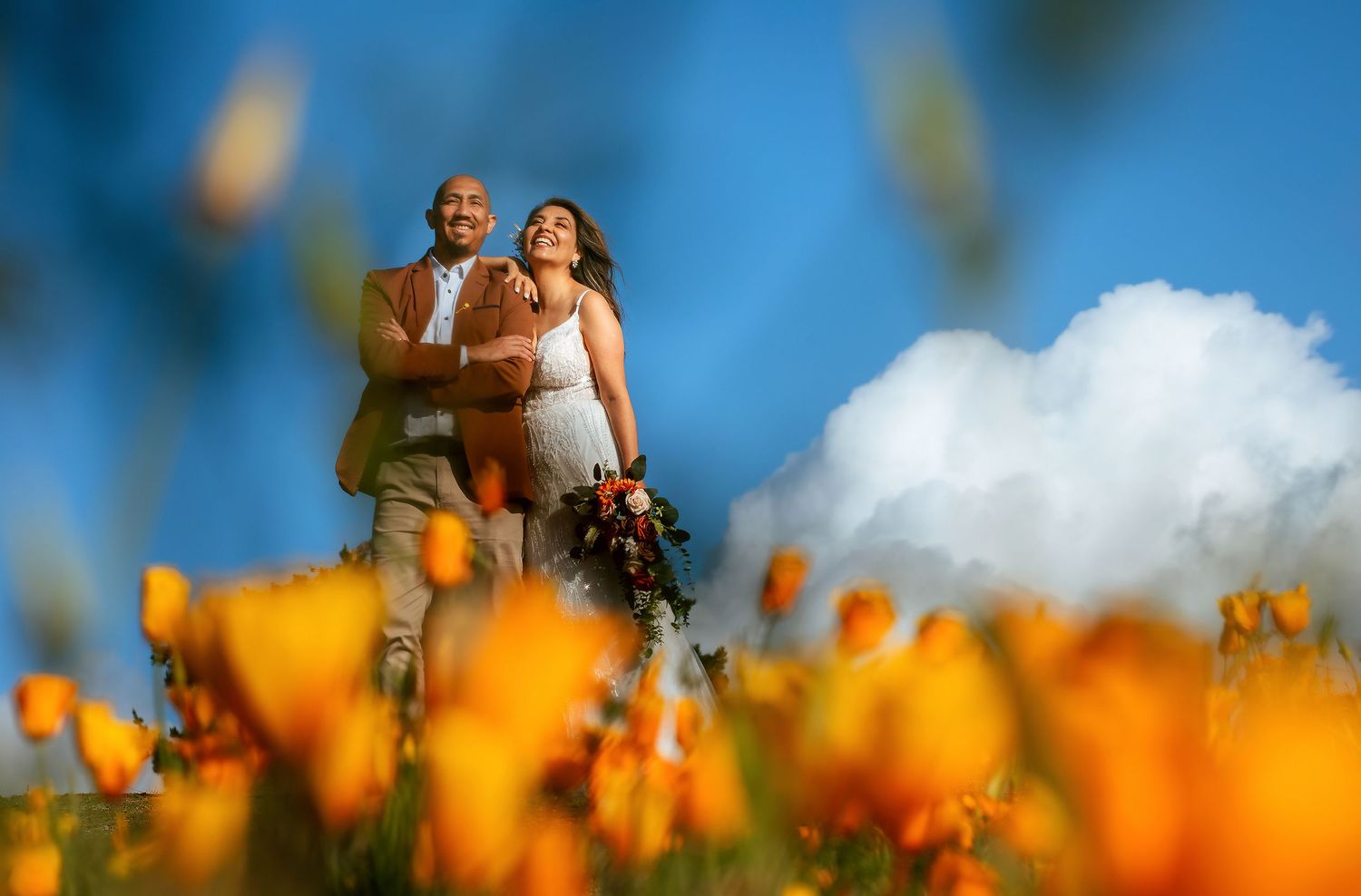 Smiling bride and groom embrace in a field of orange poppies under a bright blue sky with a fluffy white cloud.