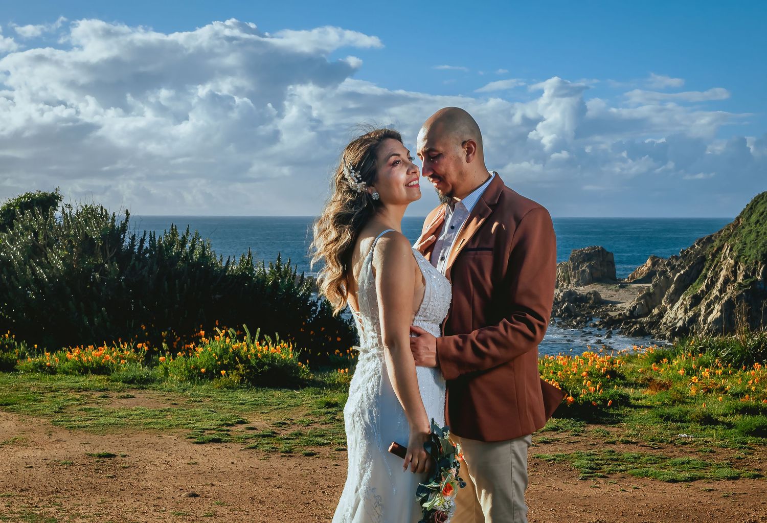 Bride in a white lace dress and groom in a rust blazer embrace on a grassy coastal cliff with wildflowers and the ocean behind them.
