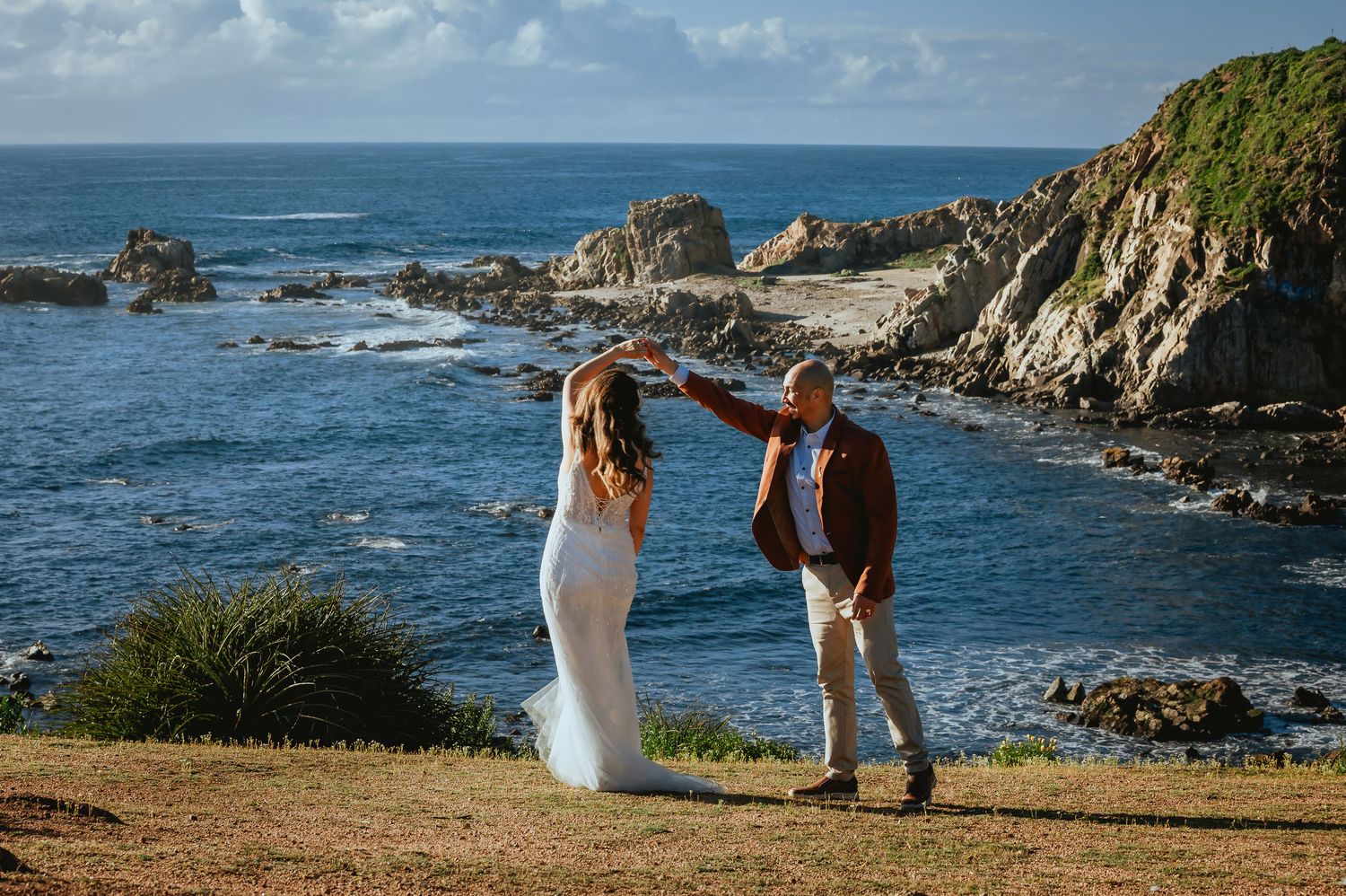Bride in a white wedding dress twirls with her partner on a grassy clifftop overlooking the ocean and rocky coastline.