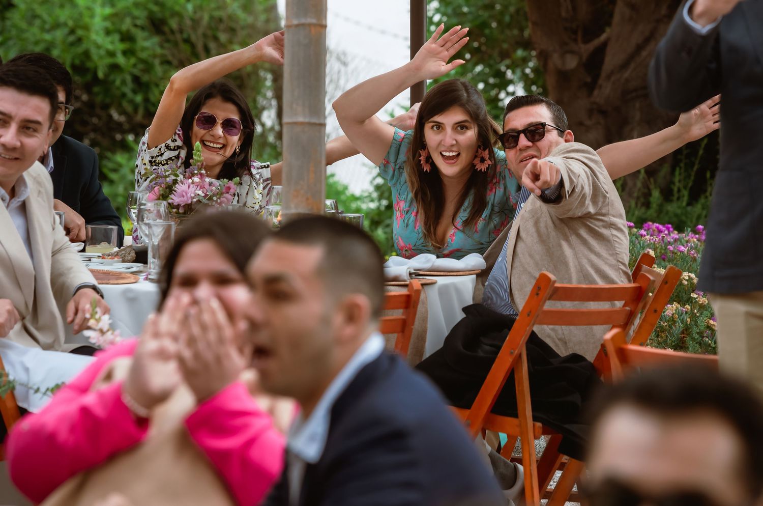 Joyful wedding guests laugh and gesture excitedly at an outdoor reception dinner table.