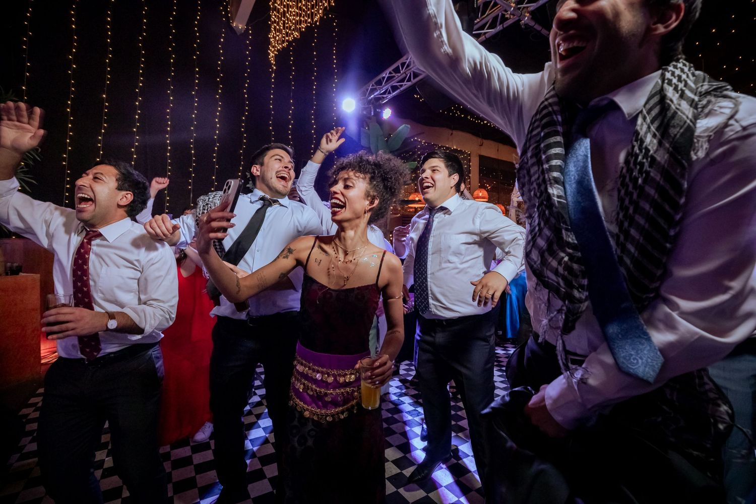 Mixed group of guests dance joyfully together on checkered floor at lively wedding reception.