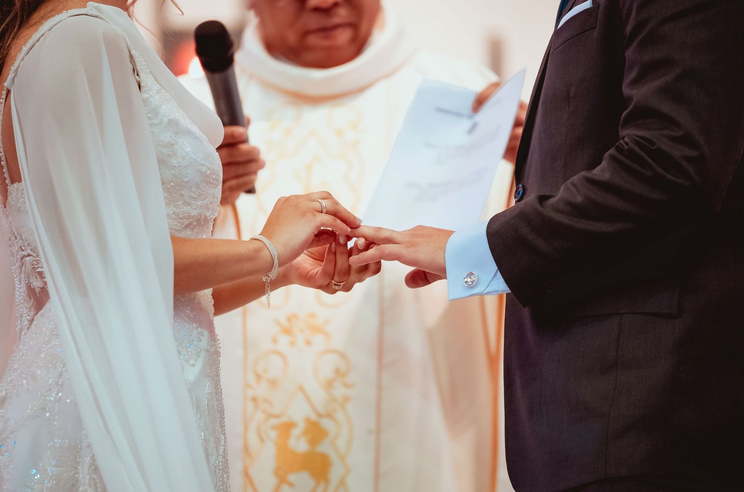 Close-up of bride placing wedding ring on grooms finger during church ceremony with priest officiating.