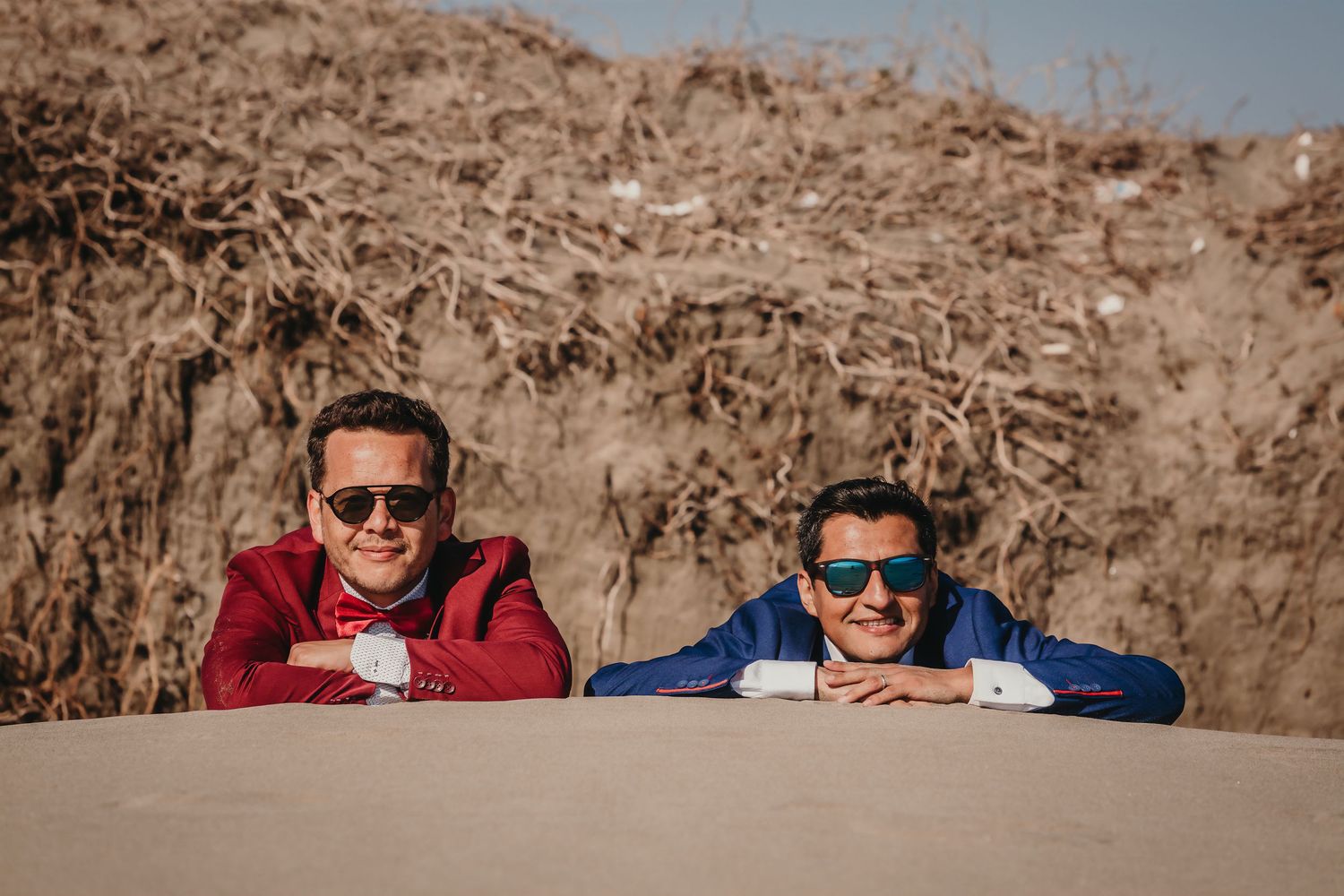 Two grooms in red and blue suits smiling and resting chins on hands against a rustic backdrop.