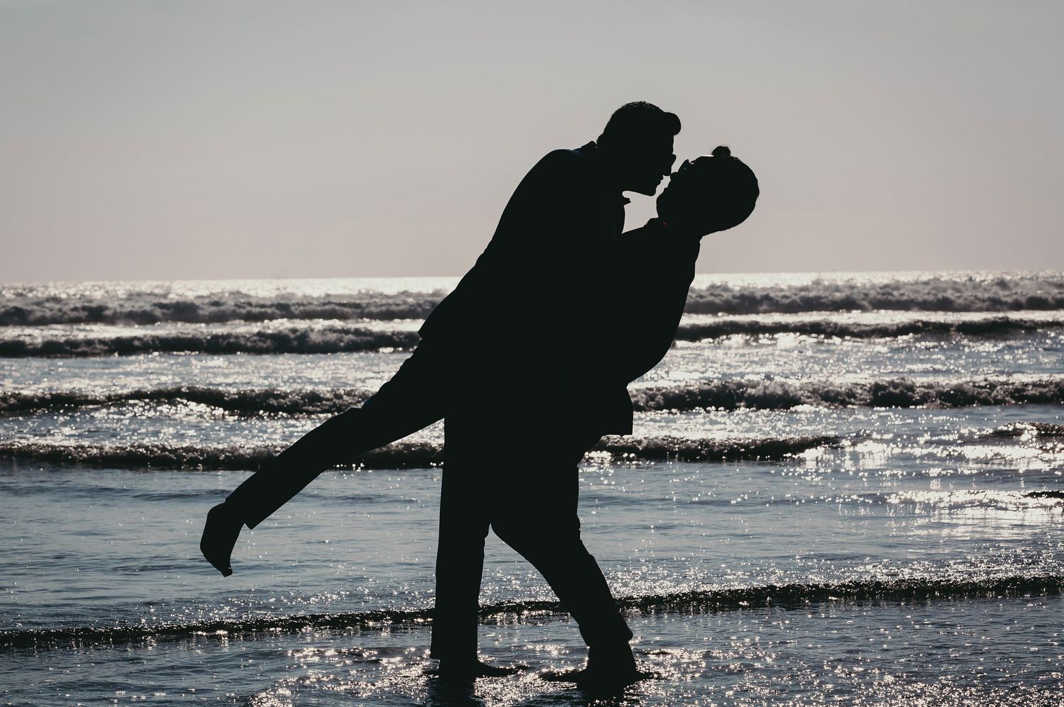 Romantic silhouette of one groom lifting his partner for a kiss on a beach at dusk.