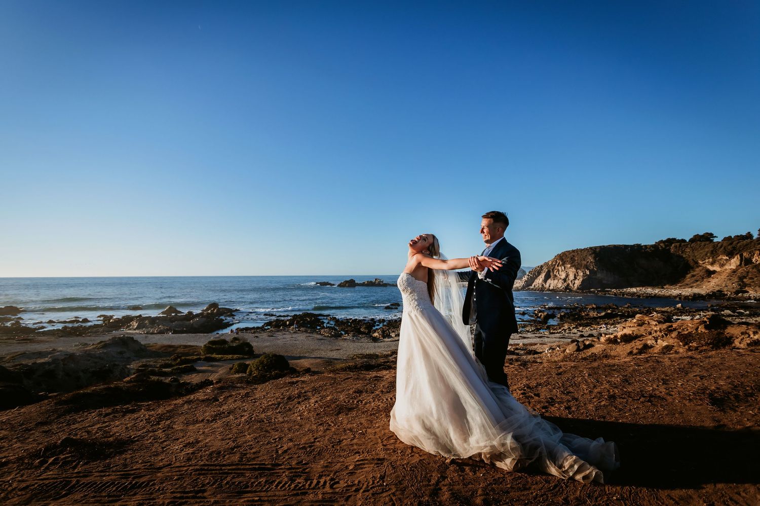 Bride and groom dance together on dramatic rocky coastal bluff with ocean horizon at golden hour.