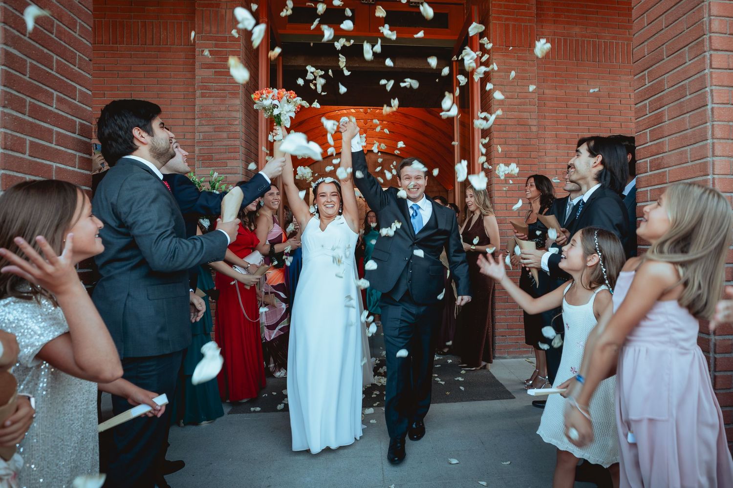 Bride and groom exit brick church amid confetti shower from celebrating wedding guests.