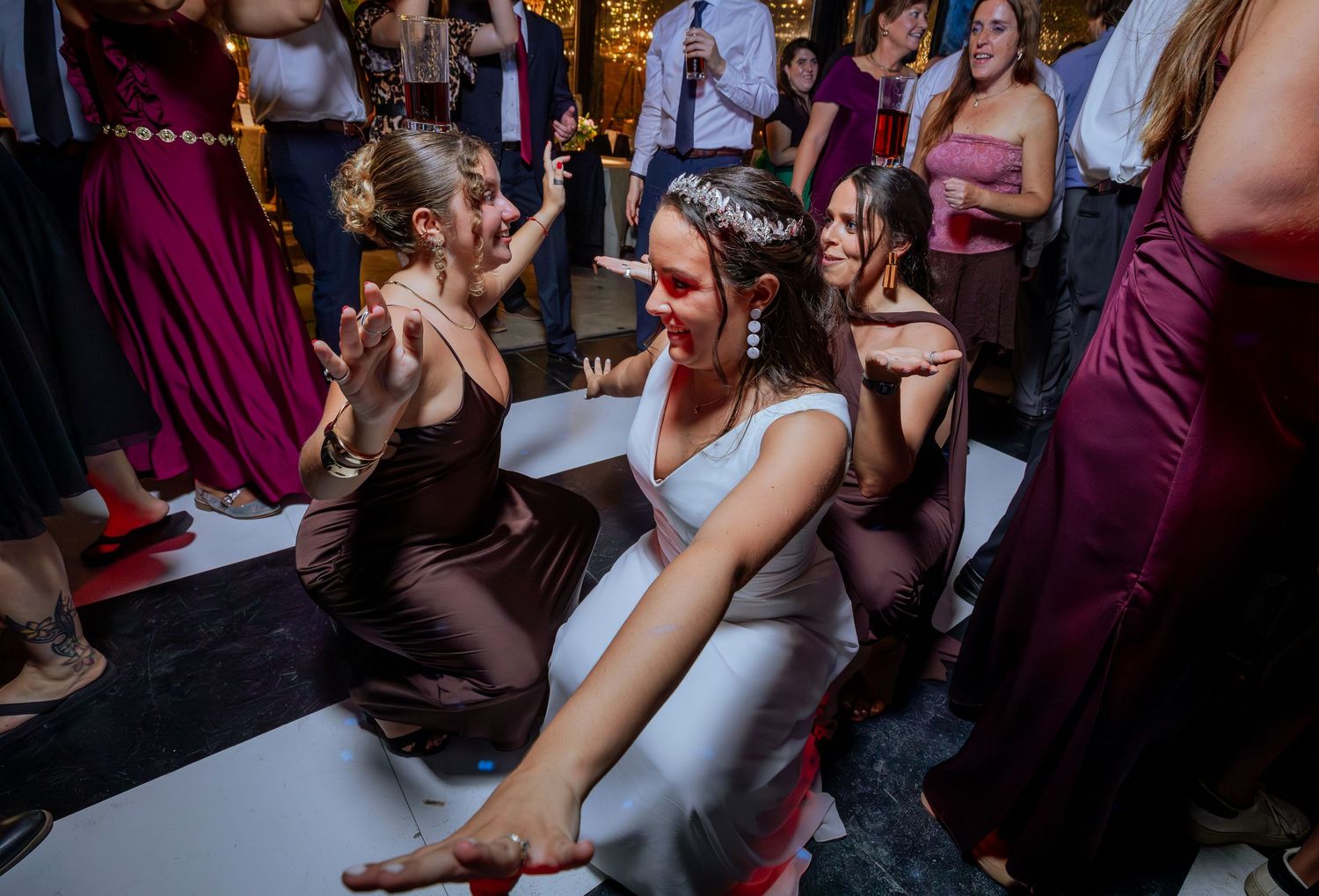 Bride in white gown laughing and dancing on the floor with bridesmaids in burgundy dresses.