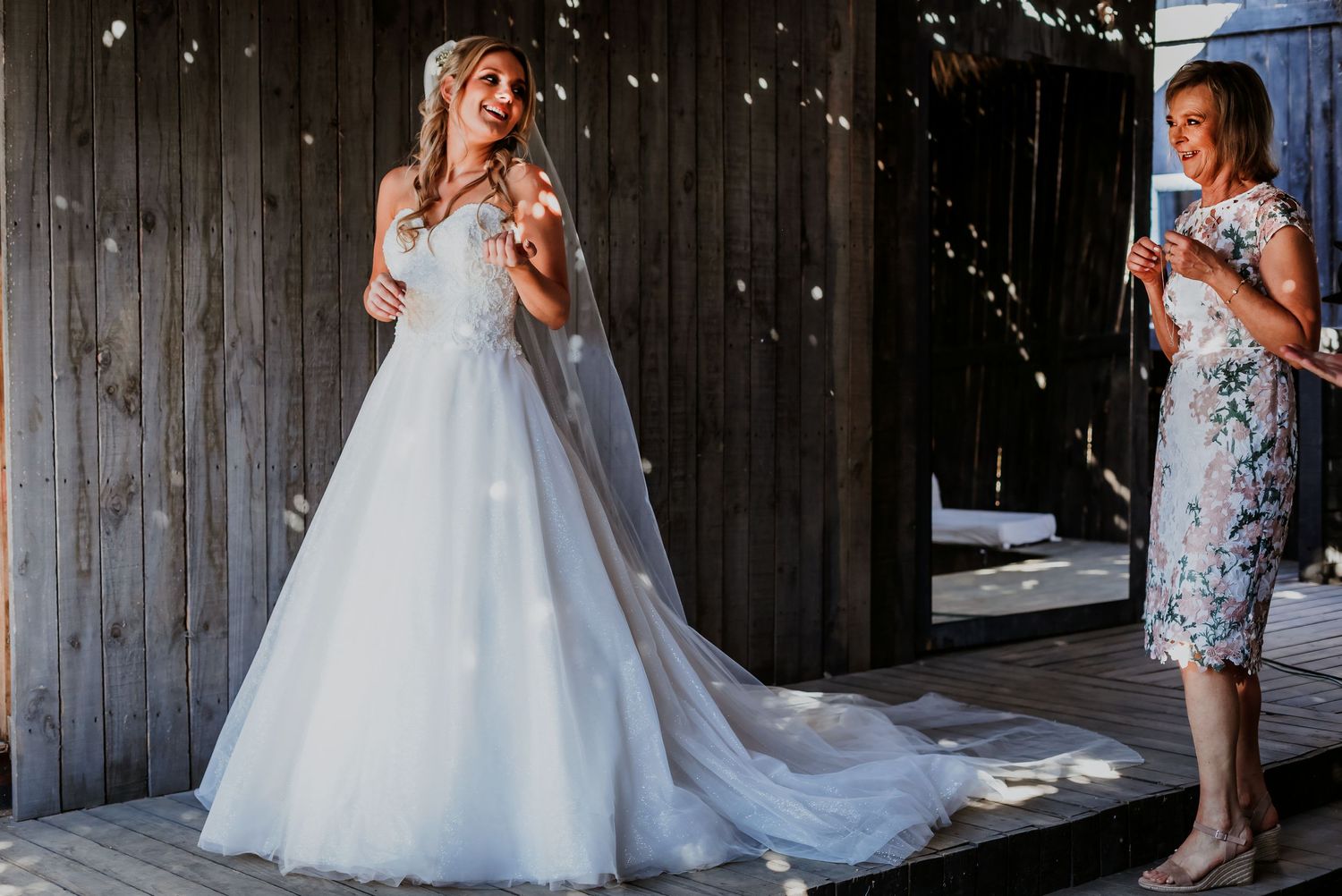 Bride in white ball gown laughing joyfully while woman in floral dress looks on at rustic barn wedding.
