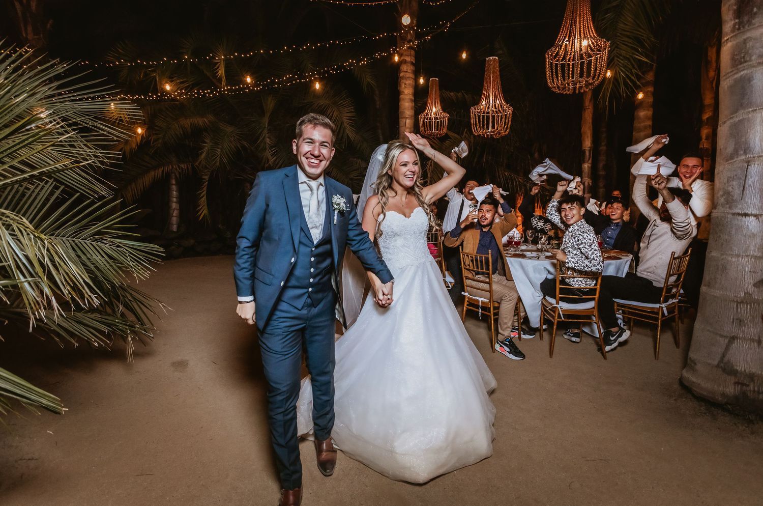 Bride and groom make grand entrance at outdoor wedding reception with string lights and chandeliers.
