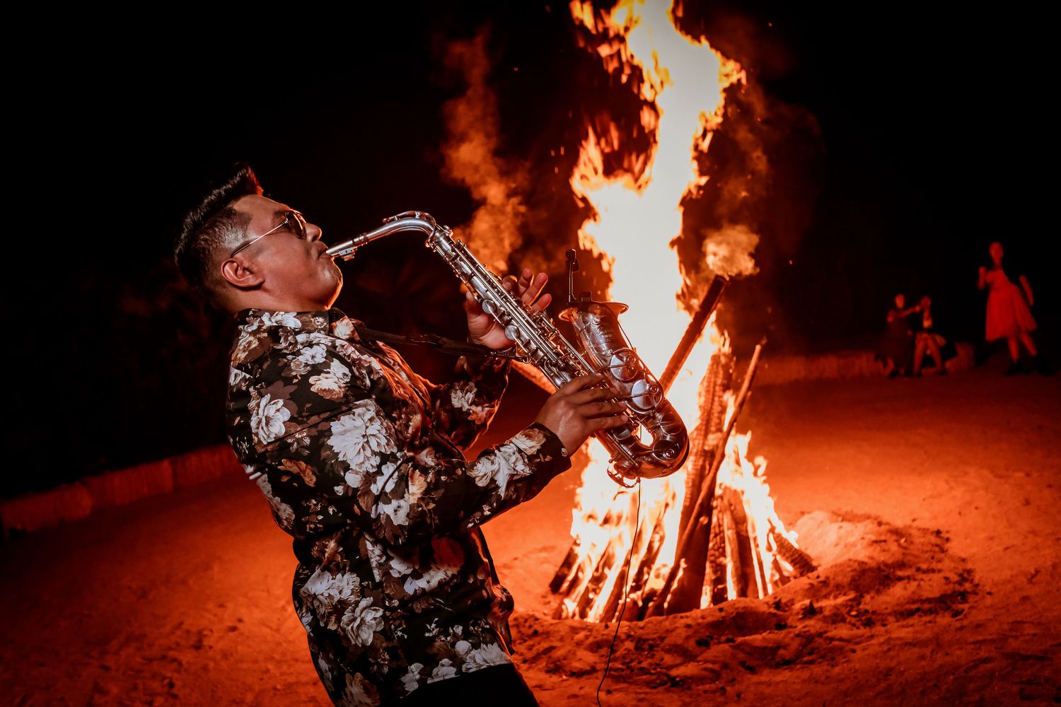Saxophonist in floral shirt performing passionately in front of a large bonfire at an outdoor wedding.