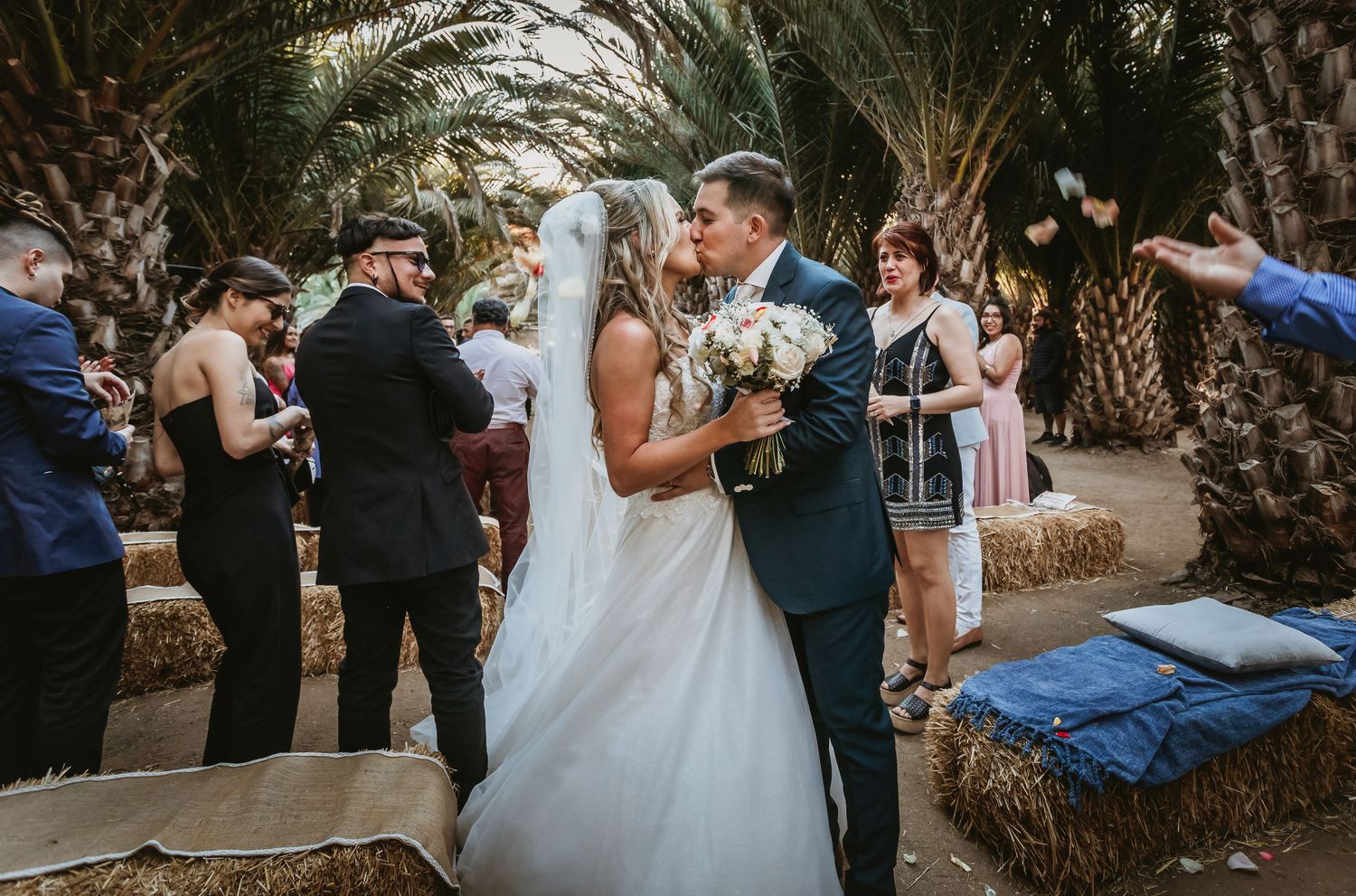 Newlyweds share first kiss at outdoor wedding ceremony surrounded by guests and palm trees.