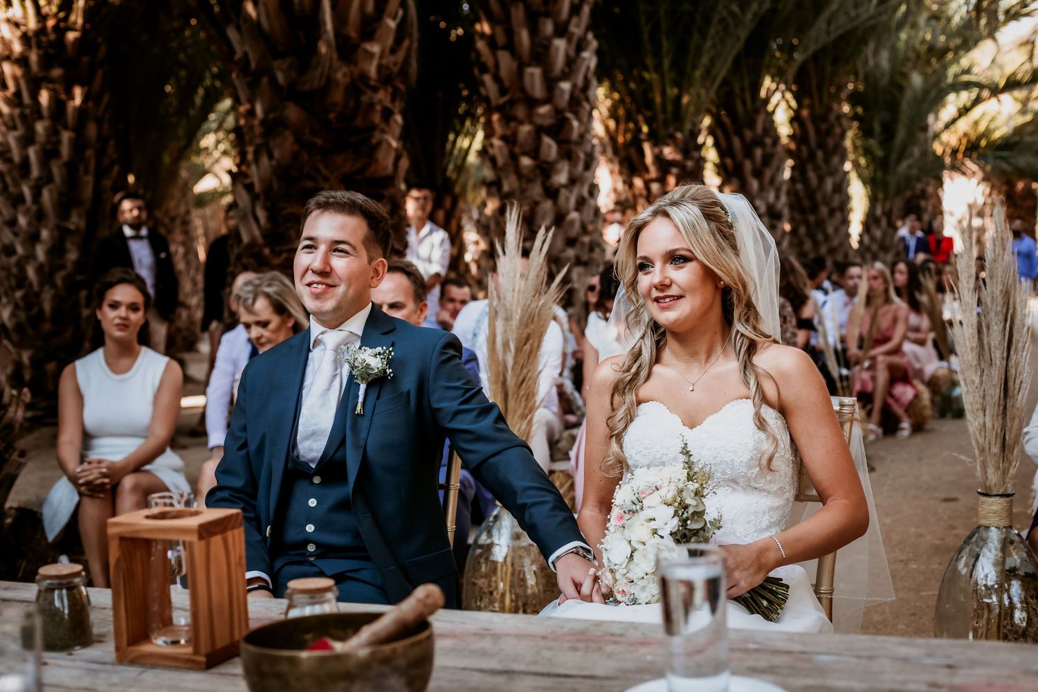 Smiling bride and groom hold hands at altar during outdoor boho wedding ceremony with guests.