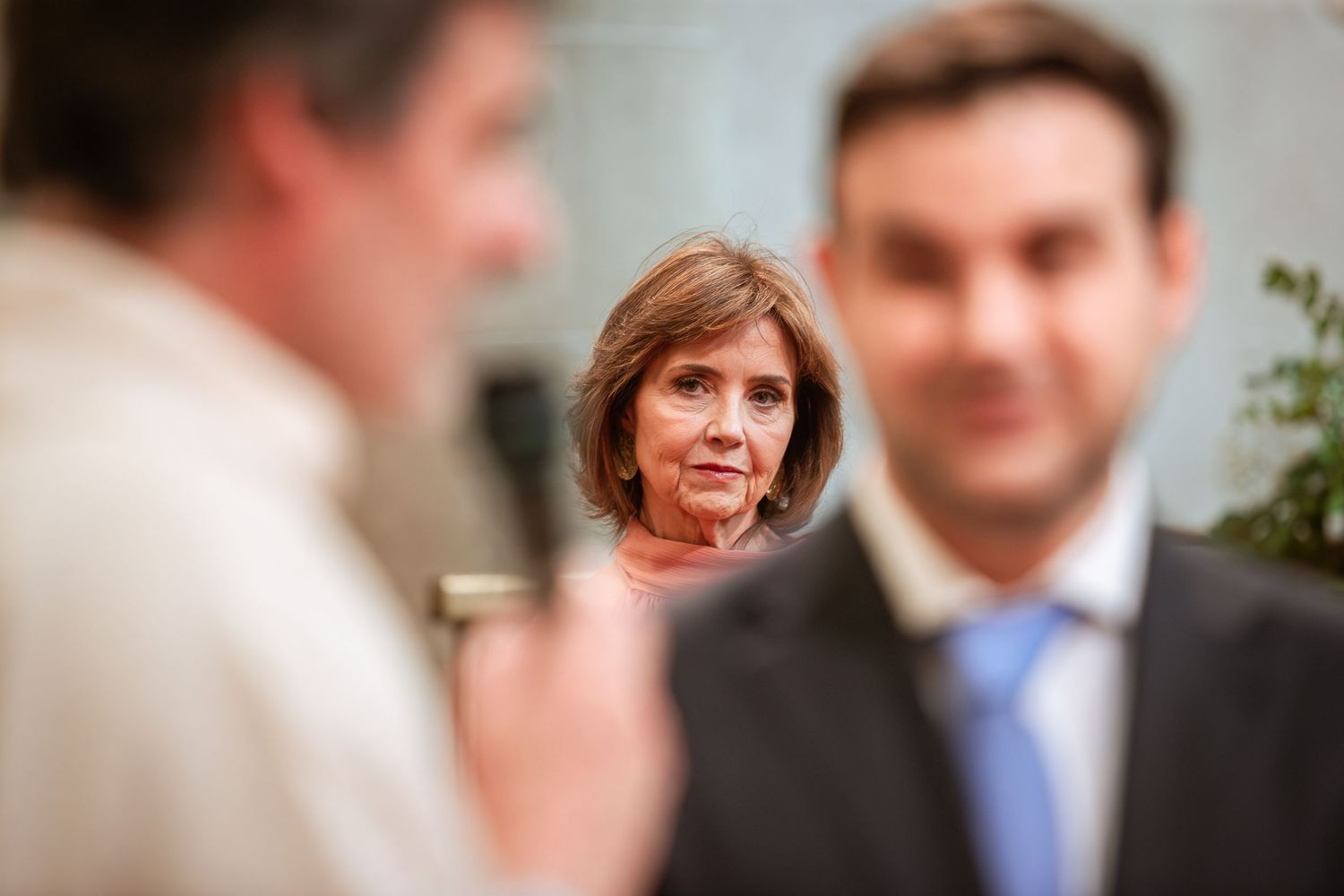 Woman in formal attire observes wedding ceremony with man in suit in foreground.