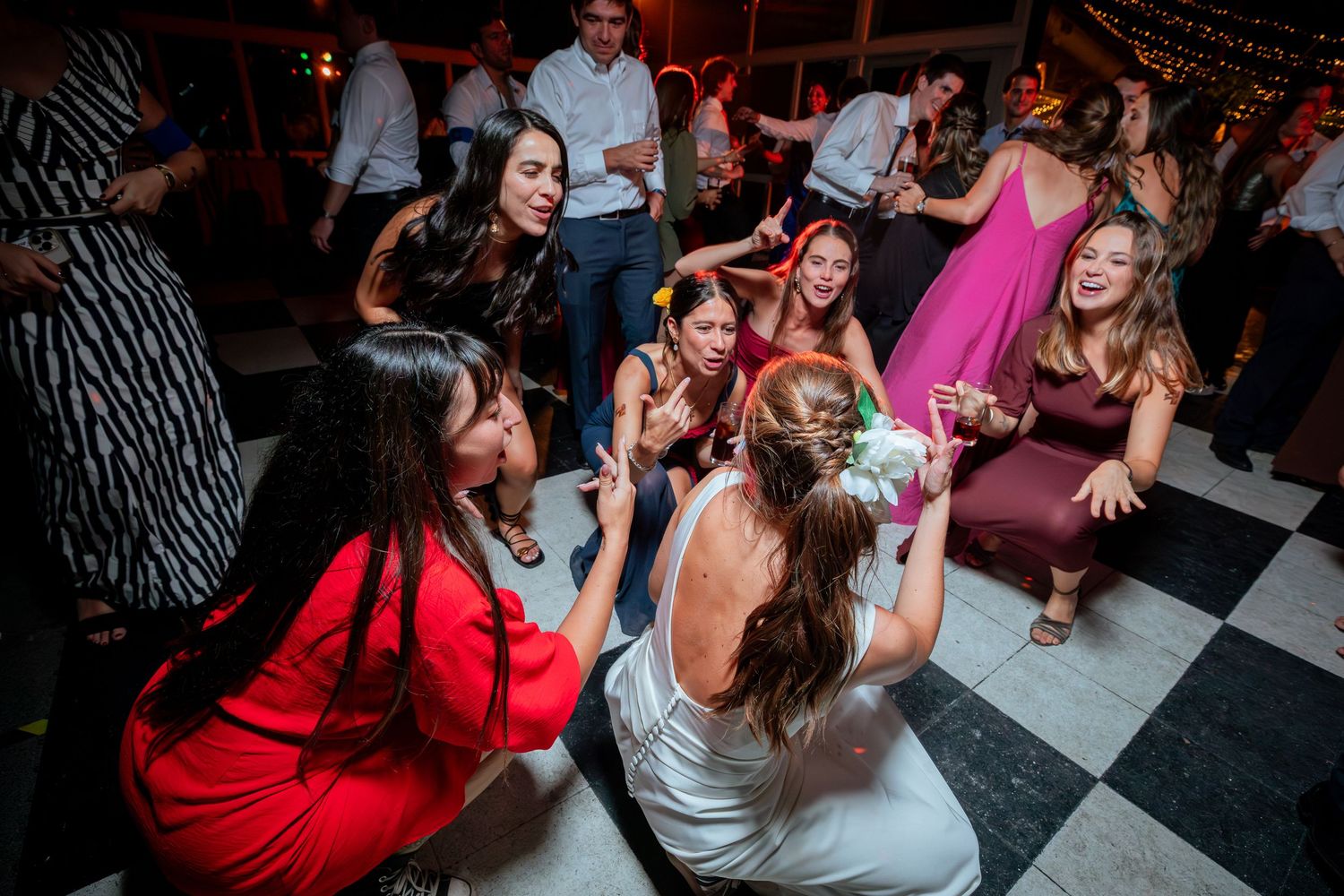 Bride crouches on checkered dance floor surrounded by excited wedding guests at lively reception party.
