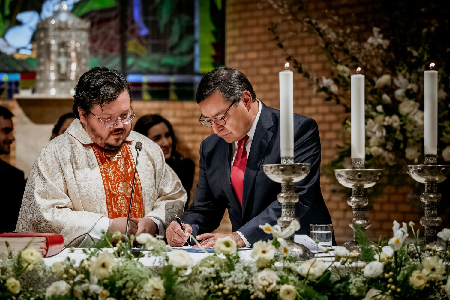 Two men signing wedding documents at a decorated altar with candles and white floral arrangements in a church.