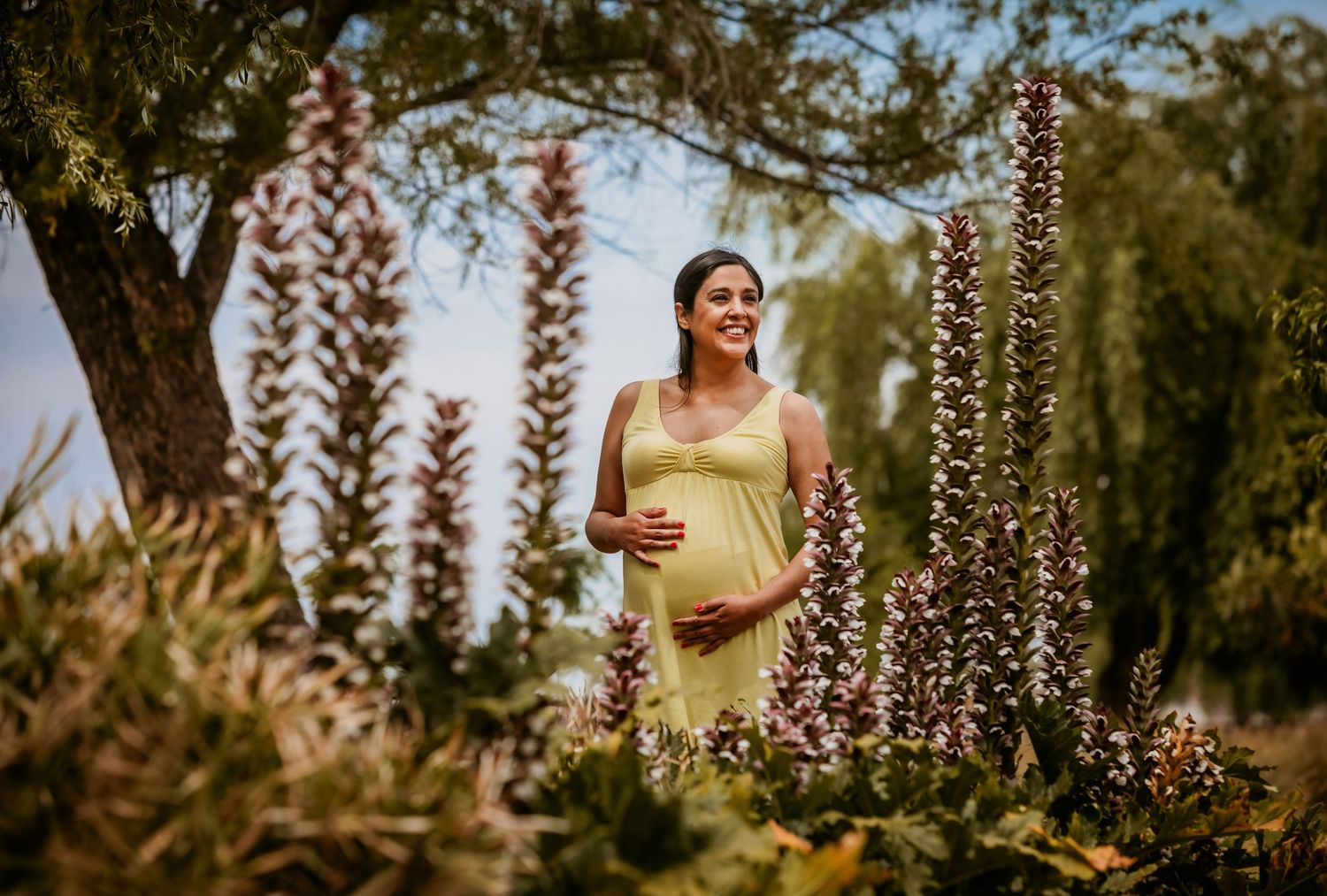 Smiling pregnant woman in yellow dress standing among tall flowering plants in a lush garden setting.