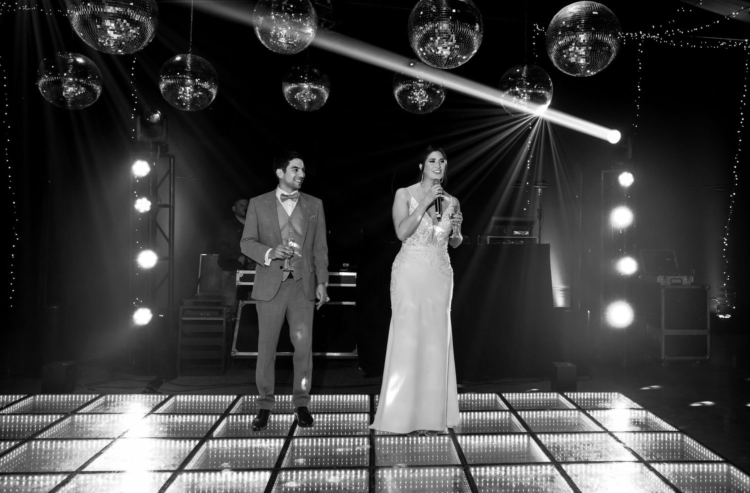 Black and white photo of a couple in formal attire standing on a dance floor under disco balls and stage lights.
