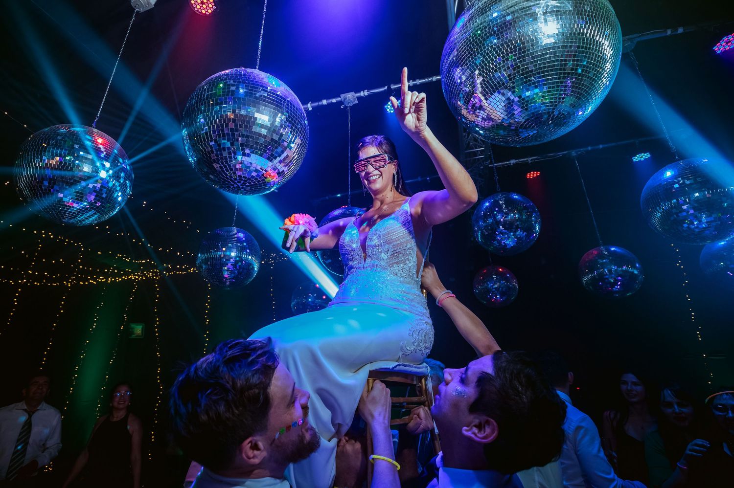 Bride lifted by guests at a festive wedding reception with glittering disco balls and colorful lights.