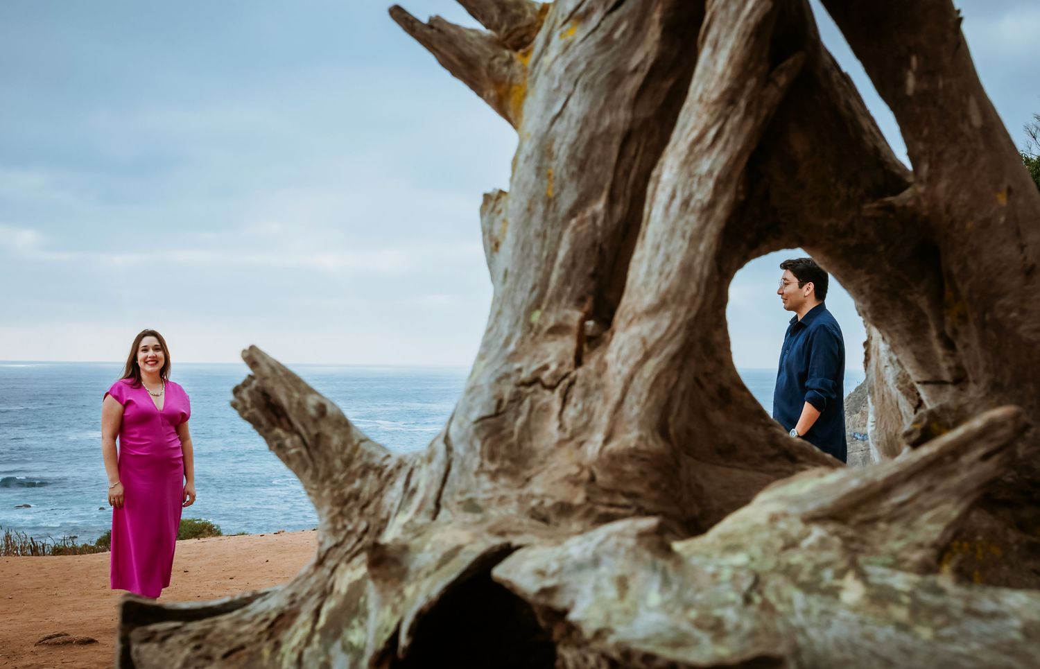 Couple poses near a large driftwood sculpture on a coastal cliff with ocean views in the background.