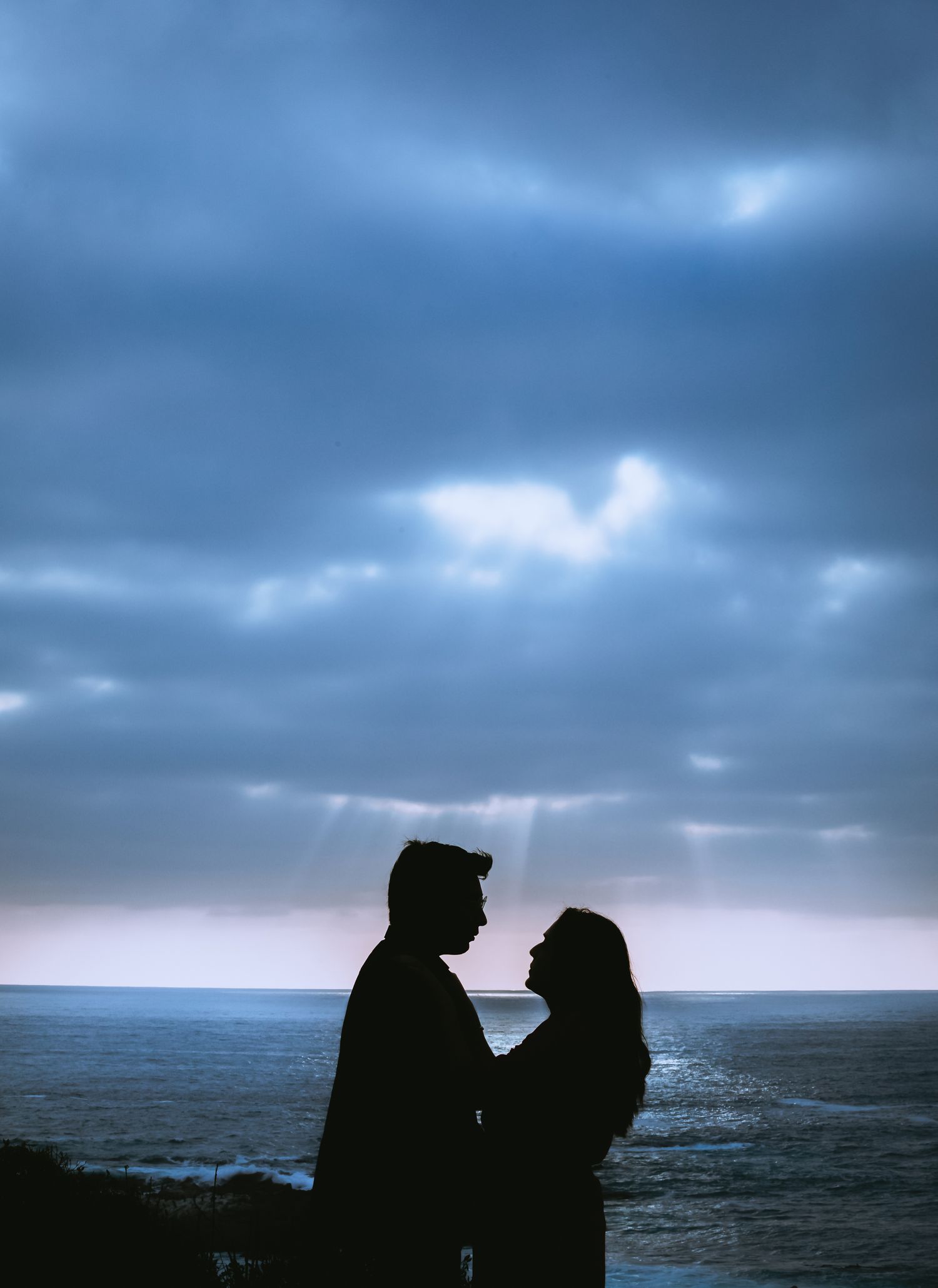 Romantic silhouette of a couple facing each other by the ocean under dramatic blue cloudy sky at dusk.