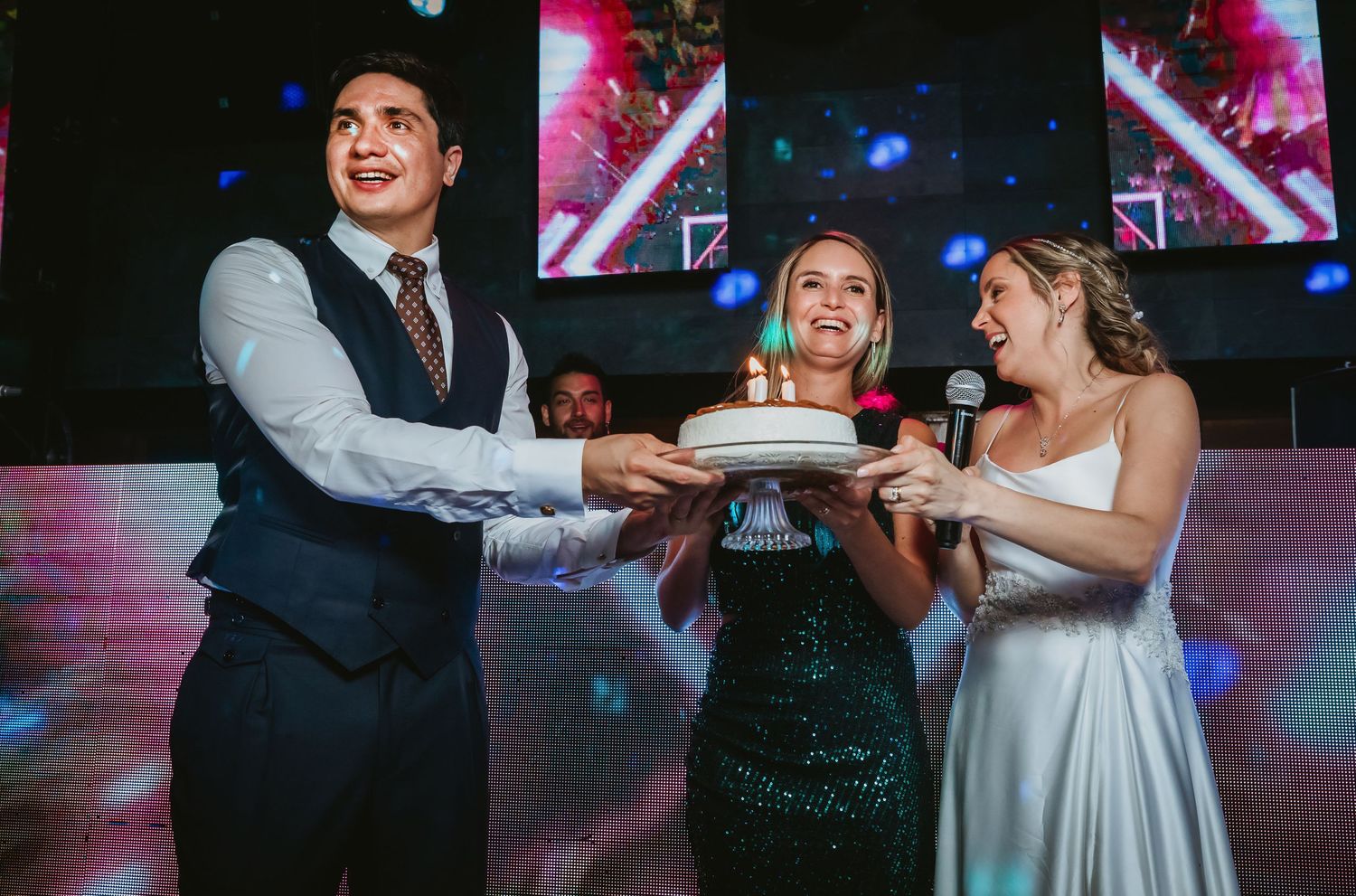 Groom and guest present birthday cake with candles to smiling bride at wedding reception.