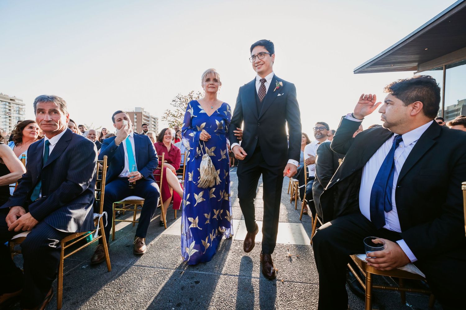 Bride in blue floral gown walks down outdoor wedding aisle with escort among seated guests.