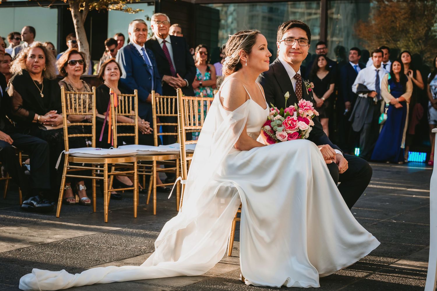 Bride in off-shoulder gown and groom sit together holding pink floral bouquet at ceremony.