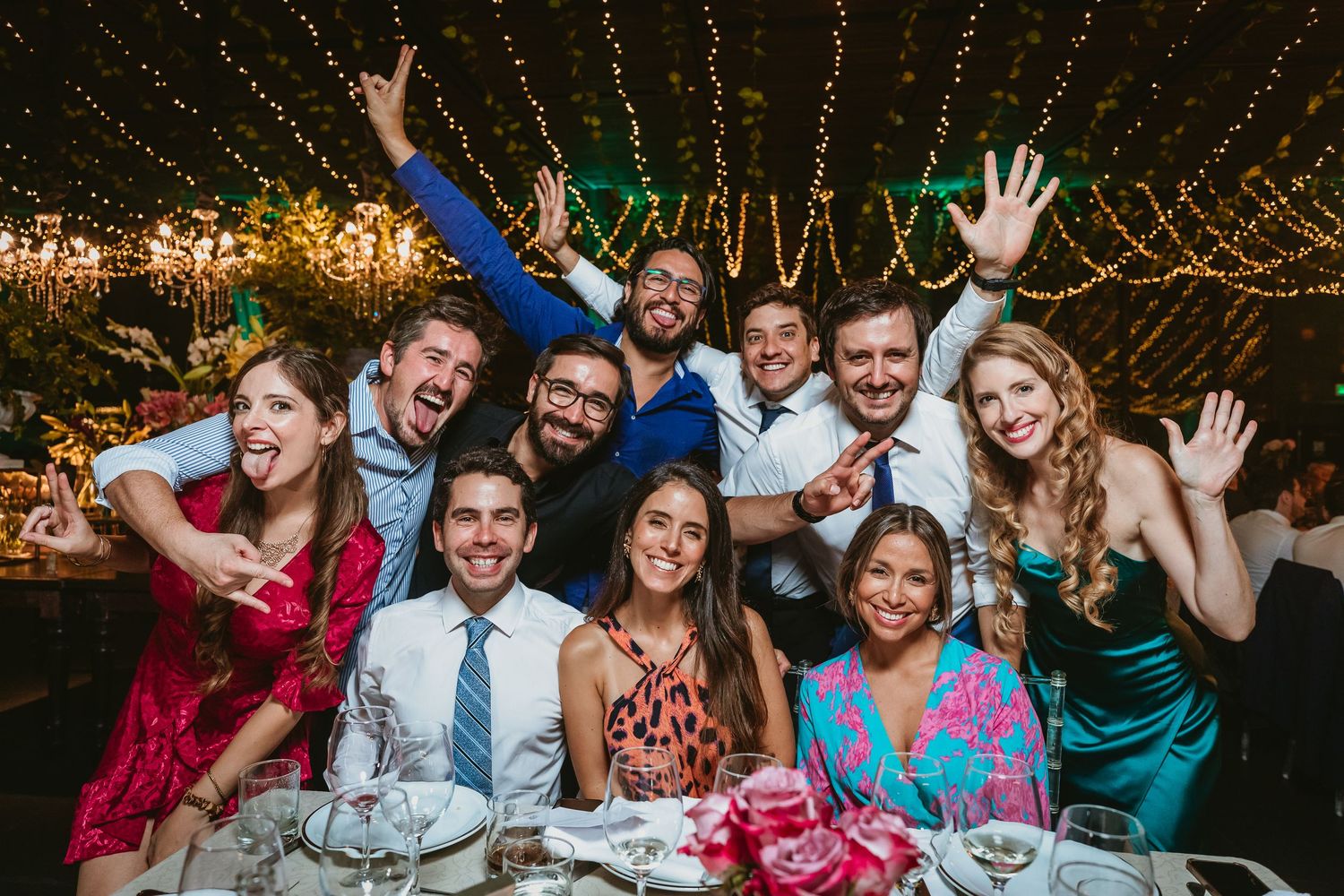 Group of friends smiling and waving at a festive wedding reception dinner under string lights.