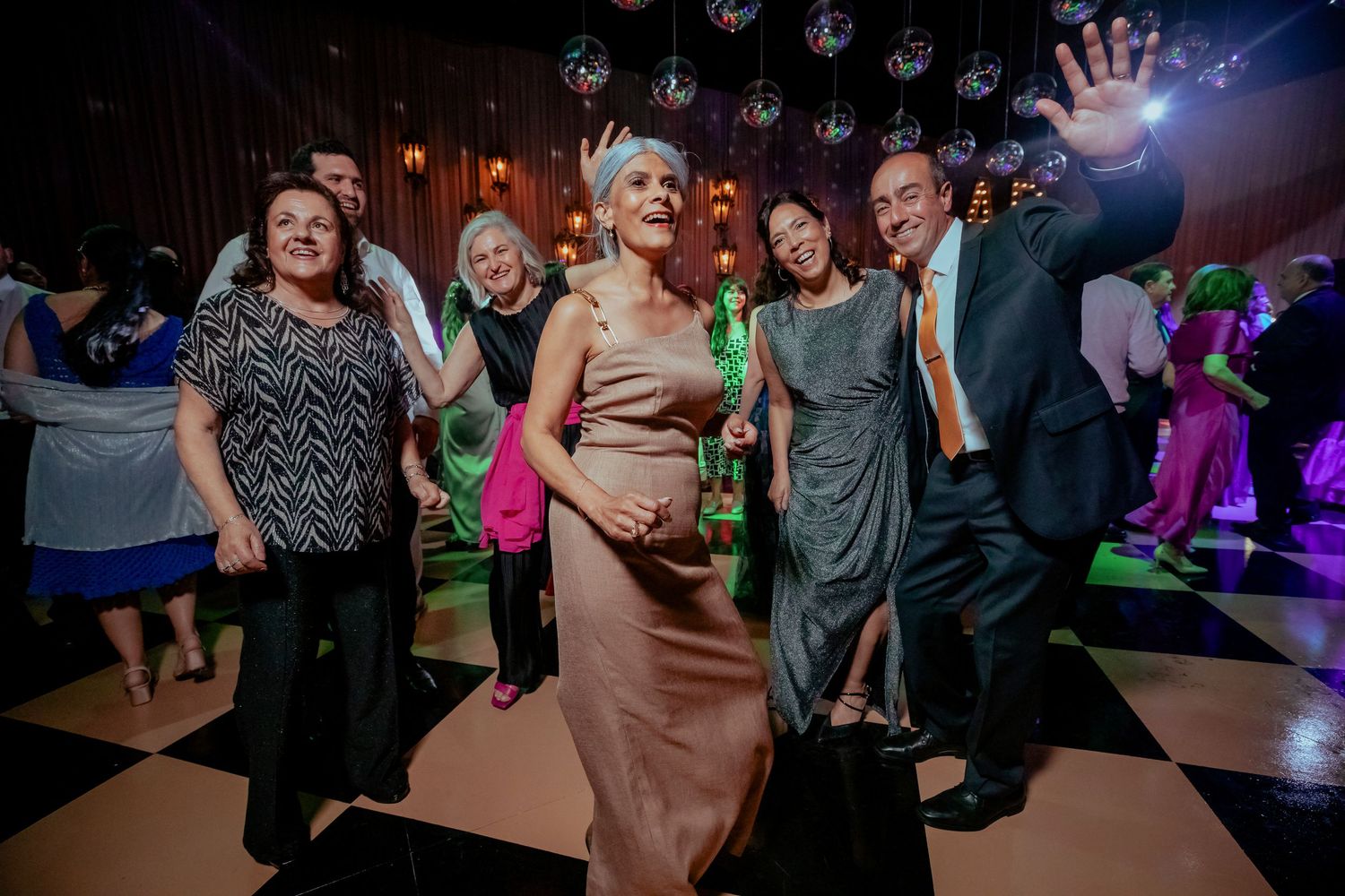 Wedding guests dancing joyfully on checkered dance floor with disco balls hanging overhead at elegant reception.
