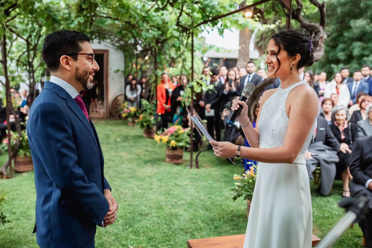 Bride reads vows to smiling groom at outdoor wedding ceremony surrounded by guests.