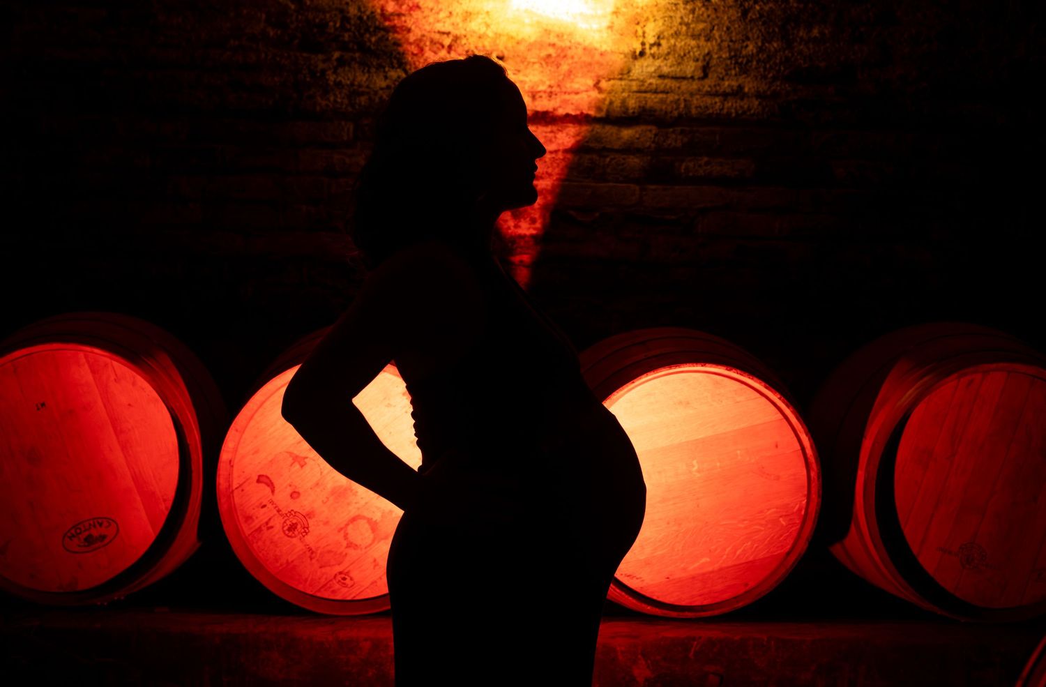Silhouette of pregnant woman posed against glowing red wine barrels in dimly lit cellar.